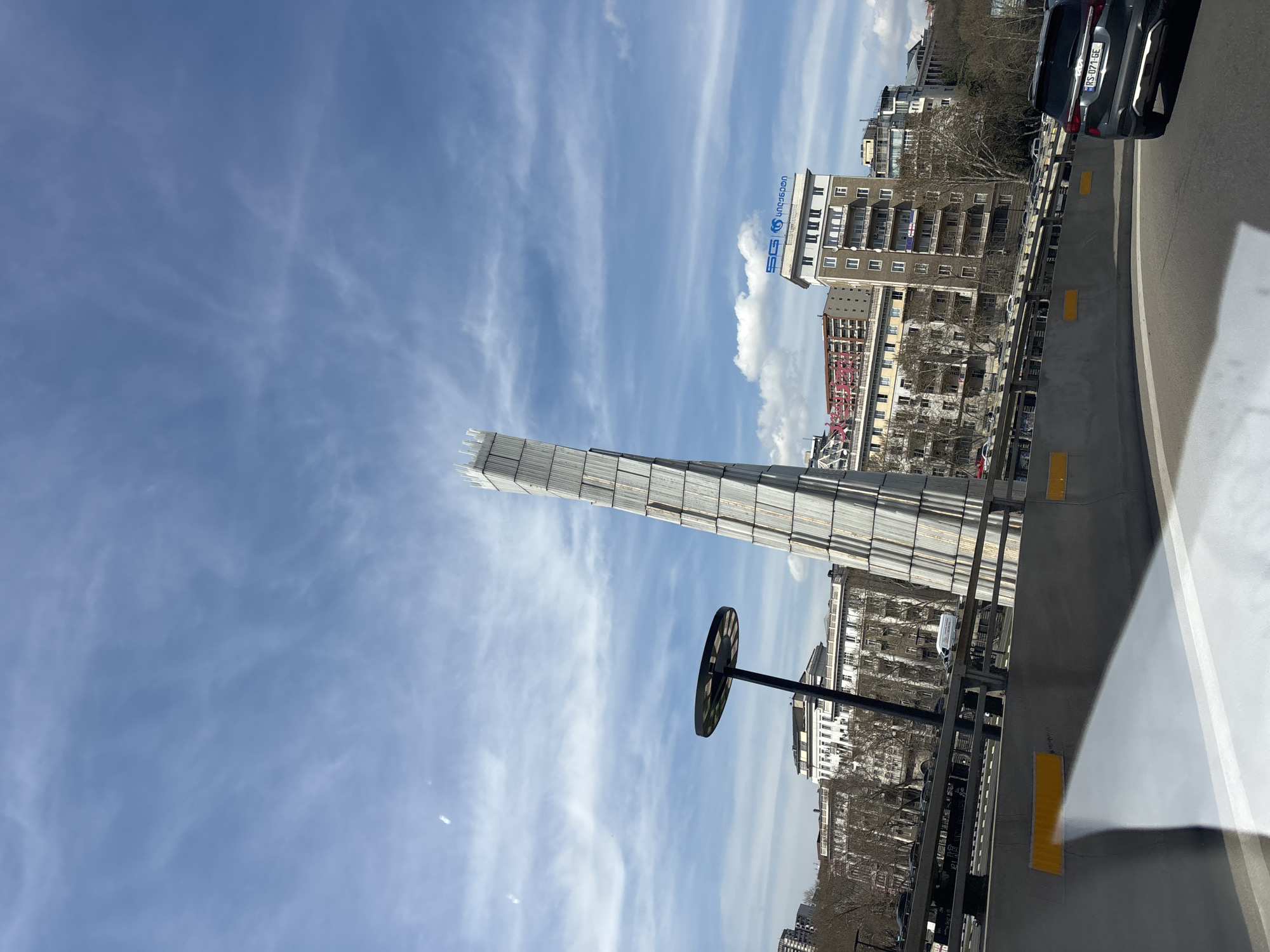 The image shows a tall, thin monument encased in scaffolding against a blue sky with scattered clouds. The scene is likely located in Tbilisi, Georgia based on the city buildings visible in the background. A black, disc-shaped object with radial markings is mounted on a pole in the foreground. The view is from a moving vehicle, as evidenced by the road barrier and passing car partially visible on the right side of the frame. A building with a blue and white logo and lettering in what appears to be Georgian script is also visible in the background.