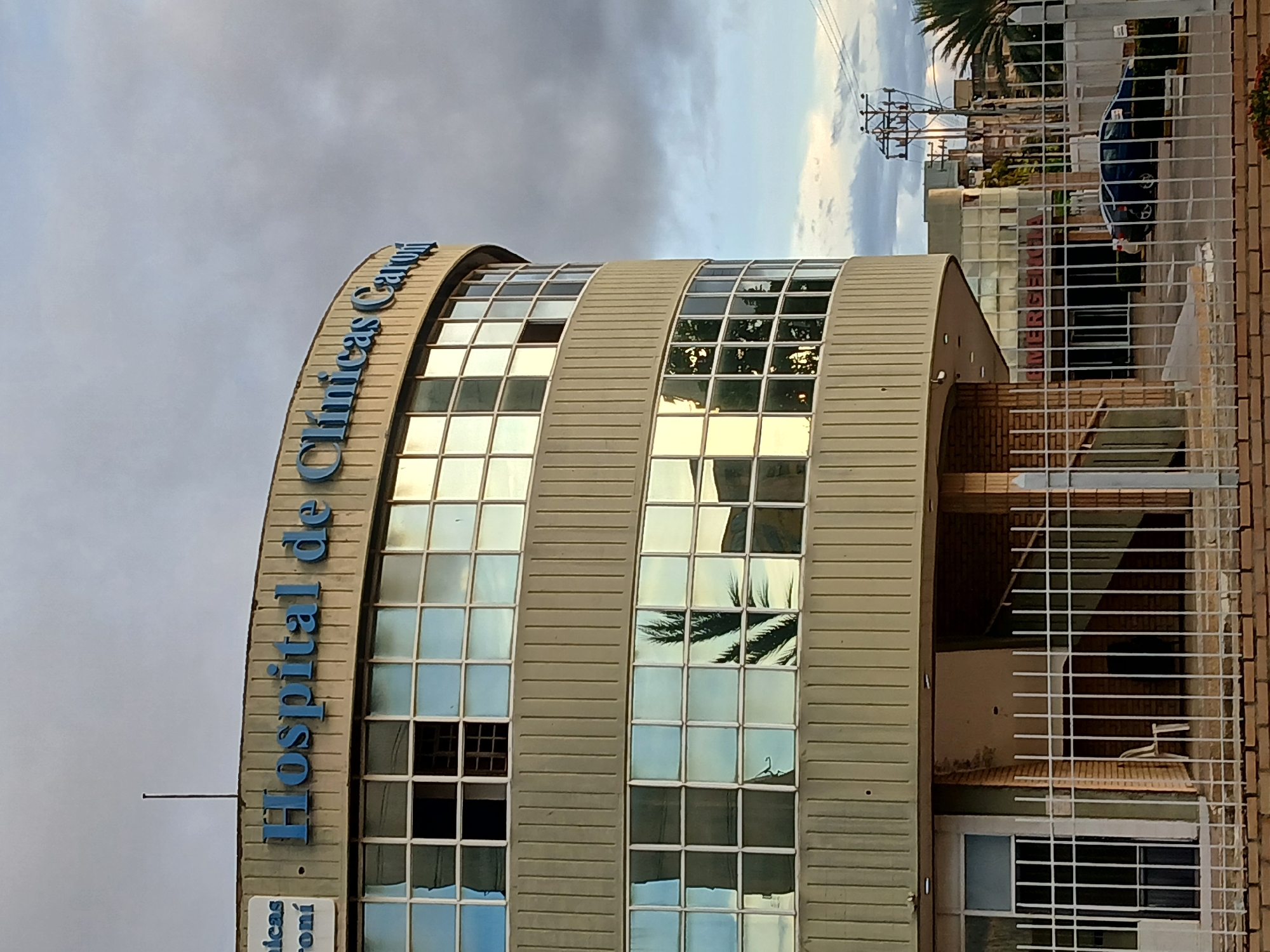 The image shows the exterior of Hospital de Clínicas Caroní in Ciudad Guayana, Venezuela. The building is a curved structure with multiple windows reflecting the sky and surrounding vegetation. The hospital's name is displayed in blue lettering on the exterior. A fence is visible in the foreground, and power lines can be seen in the background. A dark-colored vehicle is parked near the building's entrance. The sky is overcast.