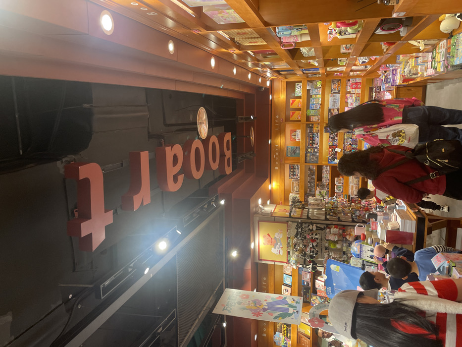 The image shows the interior of a bookstore in Chongqing, China, located at latitude 29.581, longitude 106.527. The bookstore is brightly lit with a large sign above the shelves that reads "BOART LIFE" in red lettering. The walls are a warm brown color and the shelves are filled with books, toys, and other merchandise. There are several people browsing the shelves, including a young woman in a pink and white striped shirt, a woman in a red sweater, and a man wearing a black jacket. A young boy in a blue shirt is looking at a book while a young girl in a red sweater is looking at a book. There are also a few young children playing with toys in the store. The floor is tiled and there are wooden tables and chairs in the middle of the store. The bookstore is organized with different sections for various types of books. The image captures the bustle and activity of a typical bookstore.