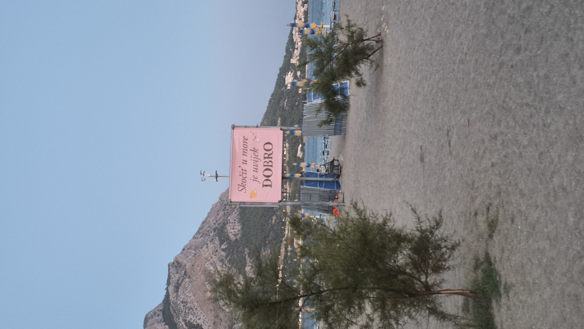 The image depicts a beach scene with a pink sign in the foreground. The sign, suspended from metal poles, displays text in Croatian. Above the text, there are two cartoonish illustrations of fish. The text reads "Skočit' u more je uvijek DOBRO," which translates to "Jumping into the sea is always GOOD." A person is seated in a beach chair behind the sign. In the background, beach umbrellas and the sea are visible, with a mountainous landscape and buildings on the coastline under a clear sky. The beach itself is sandy, with some sparse vegetation.