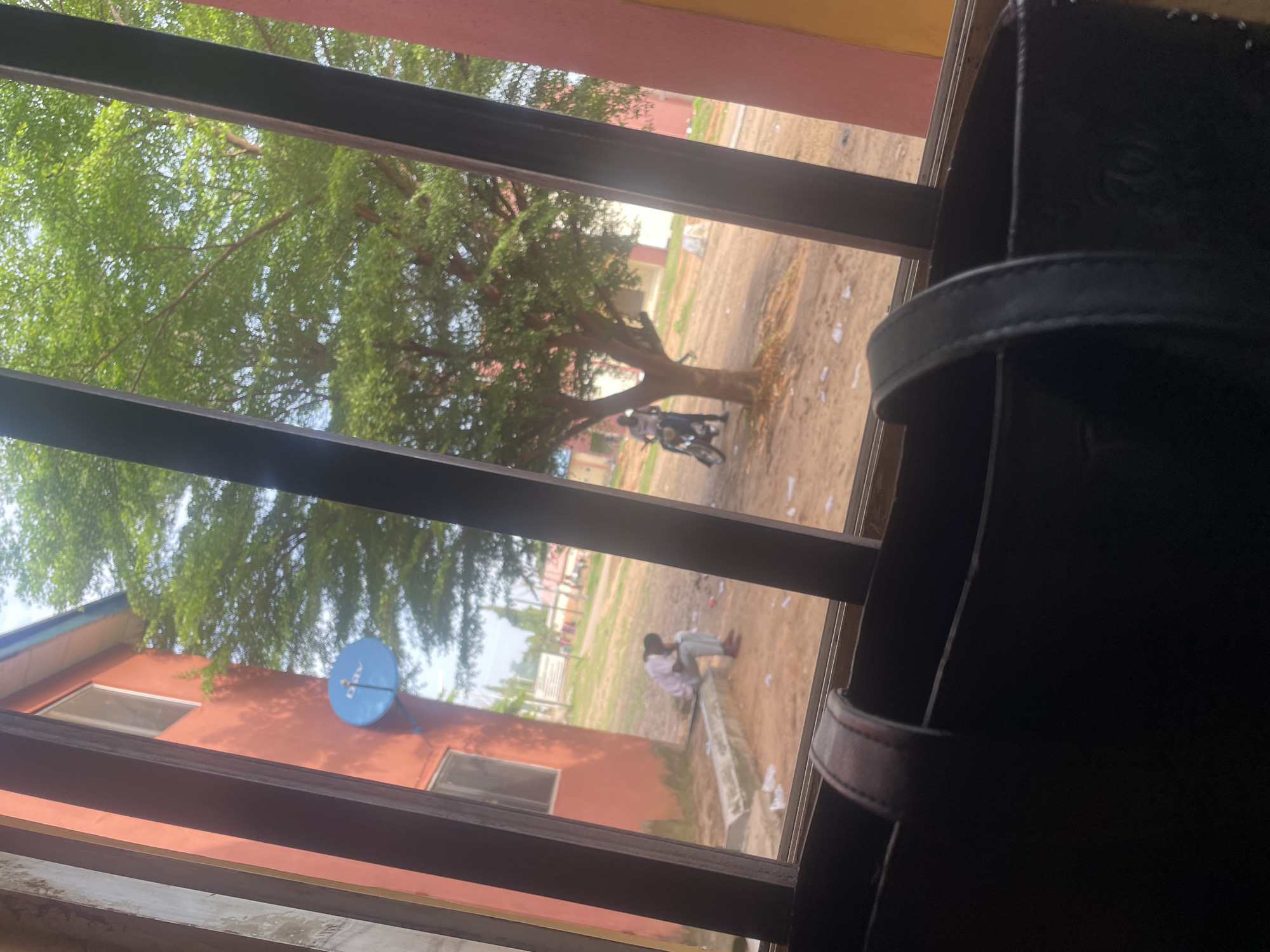 The image captures a view through a barred window, looking out onto an outdoor scene. In the foreground, a portion of a dark-colored bag with a leather strap is visible. Beyond the bars, a red building with windows is present on the left. A blue satellite dish is mounted on the side of this building.
Further into the scene, a large tree with lush green foliage dominates the middle ground. To the left of the tree, a person is seated on a concrete structure, appearing to be in a dejected pose, with their head bowed. To the right of the tree, another person is standing next to a motorcycle. The ground is sandy and somewhat littered with debris. In the background, other buildings and some distant figures can be observed, suggesting a populated area, possibly a school compound or residential area within Lokoja, Nigeria. The lighting suggests it is daytime, with the sky partially visible through the tree canopy.