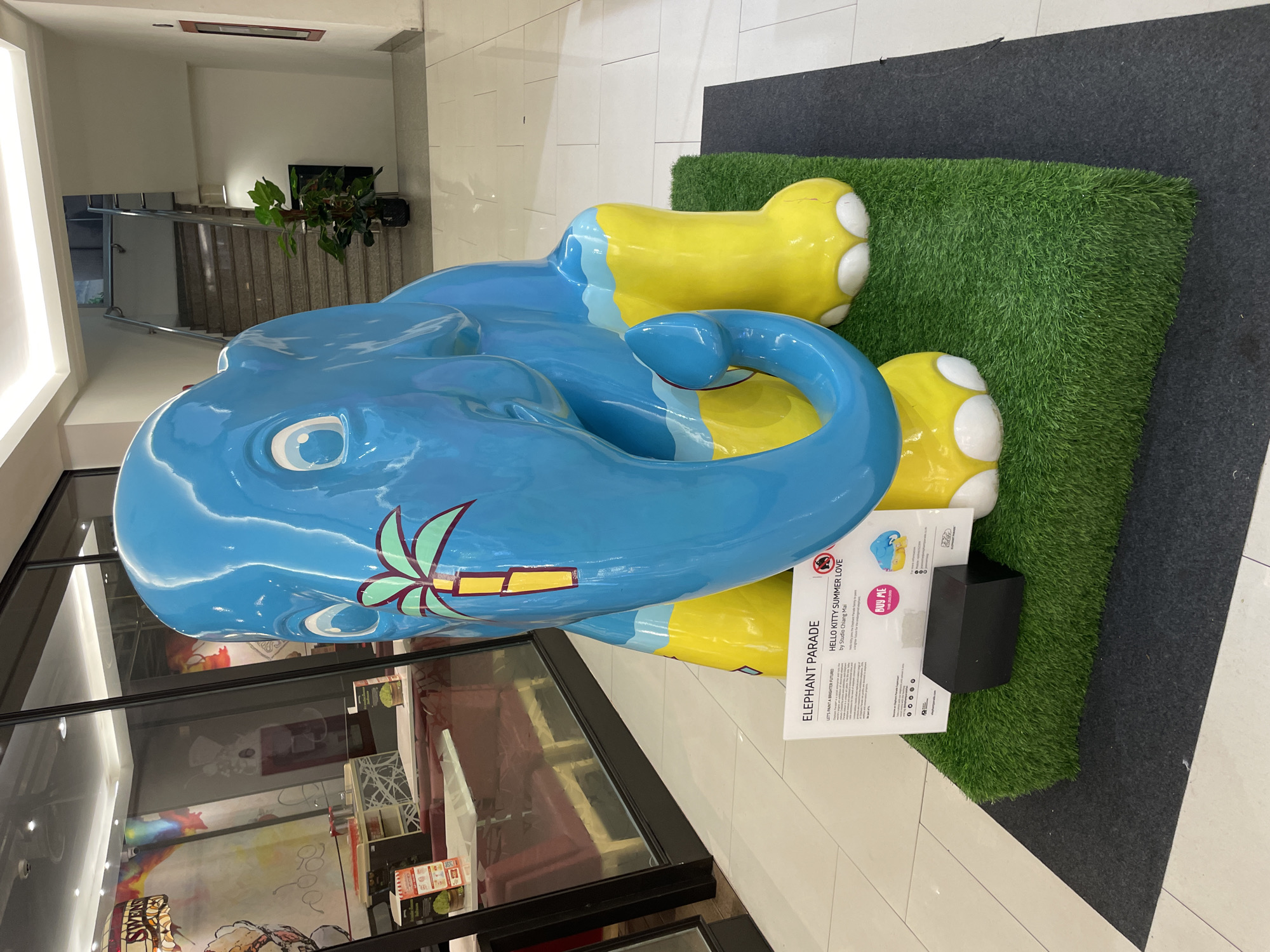 An outdoor-style fiberglass sculpture of a cartoon elephant, painted bright blue with yellow legs and white toenails, is displayed on a patch of artificial turf on a dark gray mat. The elephant has large white eyes with black pupils and a stylized green palm tree design on its forehead. A sign in front of the elephant reads "ELEPHANT PARADE" and provides details about a "HELLO KITTY SUMMER LOVE" exhibit by Studio Chiang Mai, encouraging viewers to "BUY ME" for a good cause. The elephant sculpture is situated in an indoor mall or public space, with a tiled floor visible and a reflection of a restaurant or shop with red seating booths and brightly colored art in the background. A staircase and a potted plant are visible in the background, suggesting a commercial or public indoor environment. The overall scene suggests a public art installation, possibly a charity event, in a retail or entertainment venue.