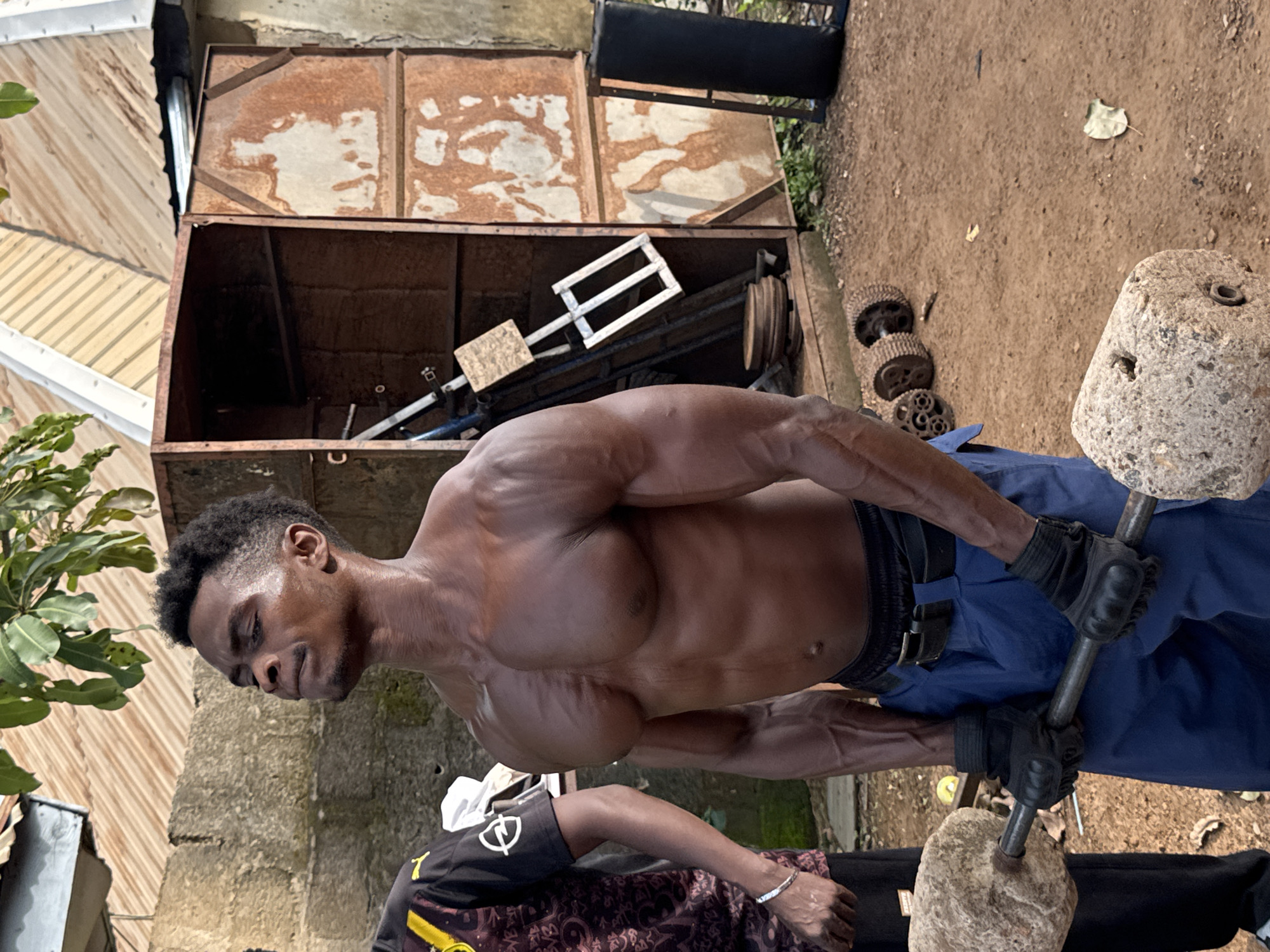 In Akwanga, Nigeria, a shirtless man with well-defined muscles is performing a strength training exercise outdoors. He is holding a barbell with what appear to be large stones as weights on either end. He is wearing blue pants and black weightlifting gloves. To his left, another person in a black and yellow shirt is partially visible. The background shows a rustic setting with a corrugated metal roof, a crumbling concrete wall, and a rusted metal cabinet containing various objects, including what looks like exercise equipment. The ground is dirt, with some leaves scattered around. The lighting suggests it is daytime.