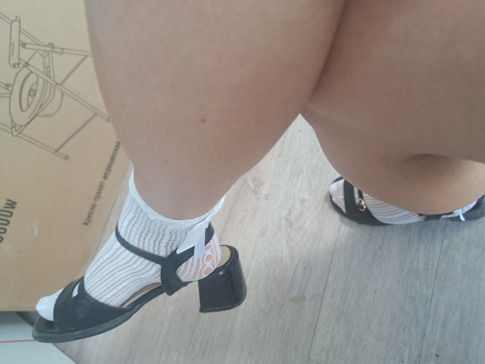 The image shows a close-up of a person's legs and feet. The person is wearing black heeled sandals with white sheer socks. The sandals have a strap across the foot with a gold buckle. The socks have a delicate lace pattern. The background is a wooden floor and a cardboard box with a drawing of a portable toilet on it. The location is Leningradskaya, Russia.