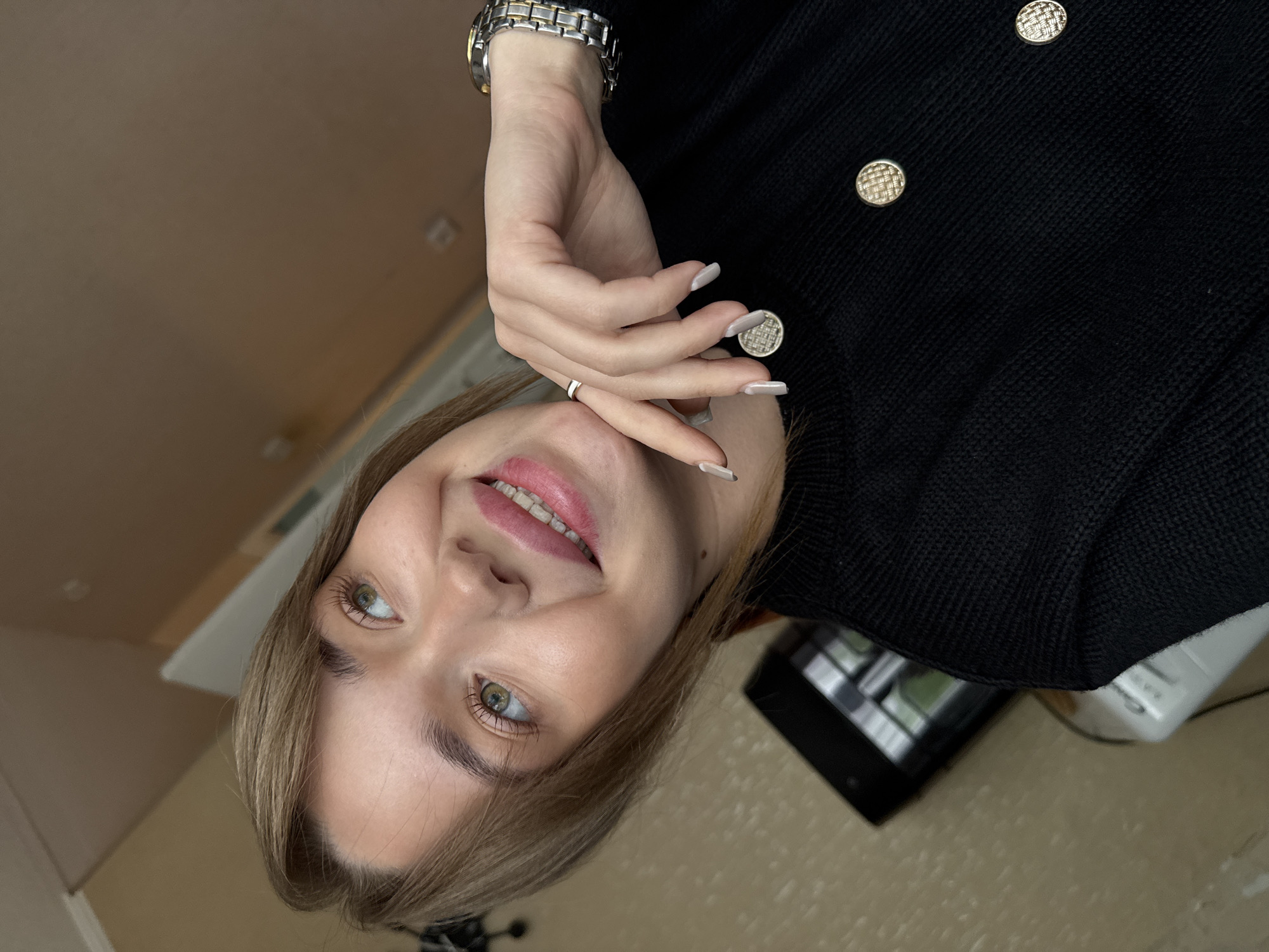 A young woman with blonde hair and green eyes, wearing a black cardigan with gold buttons and a silver wristwatch, poses for a selfie. She is smiling with pink lipstick and has a silver ring on her finger. The background shows a plain wall, and objects that suggest an indoor setting, including what appears to be electronic equipment. The location is Chaikovsky, Russia.