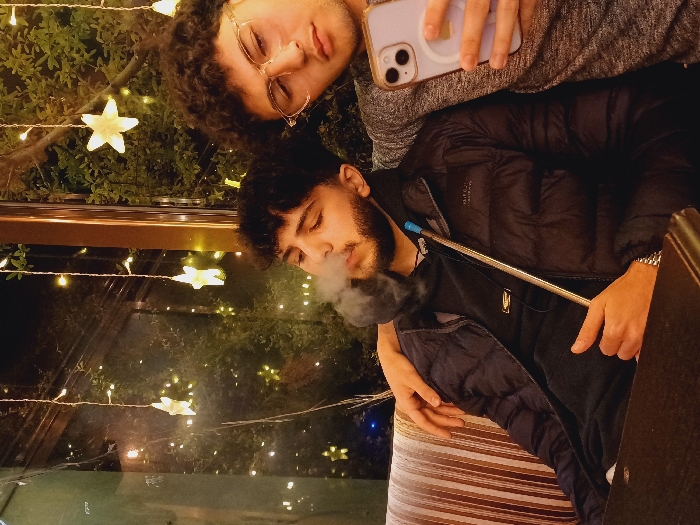 Two young men pose in a dimly lit setting adorned with star-shaped fairy lights and foliage. One, wearing glasses and a gray top, holds a phone. The other, in a black jacket, is smoking a hookah, exhaling a cloud of smoke. The image is framed within a window or glass panel, possibly at an outdoor café or lounge. The scene feels intimate and casual.