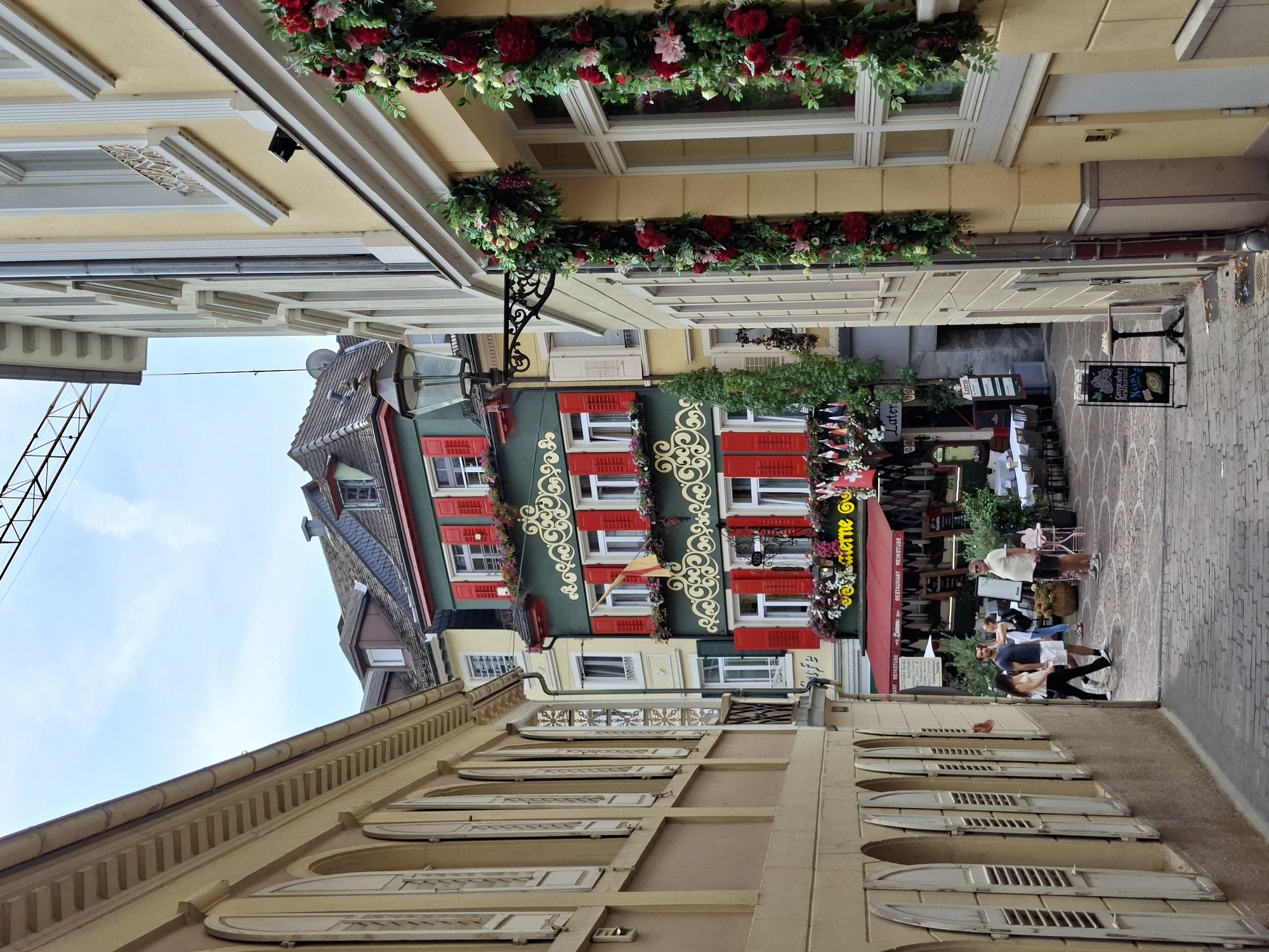 The image depicts a narrow cobblestone street in Baden-Baden, Germany, lined with historic buildings adorned with floral decorations. On the left, a building with arched windows and weathered shutters is visible. The building on the right, painted a pale yellow, features a window with a decorative grate and is draped with garlands of red and pink flowers.
Further down the street, a striking green building with red shutters and ornate yellow trim stands out. This building appears to be a restaurant or cafe, with tables set up outside under a red awning. Several people are visible in the street: a couple walking towards the viewer, a man and a woman standing near the restaurant, and another man standing further in the distance. The street is paved with cobblestones, and a black sign on a stand with chalk writing is placed on the pavement. The sky above is overcast with a hint of blue. A construction crane is visible in the upper part of the image.