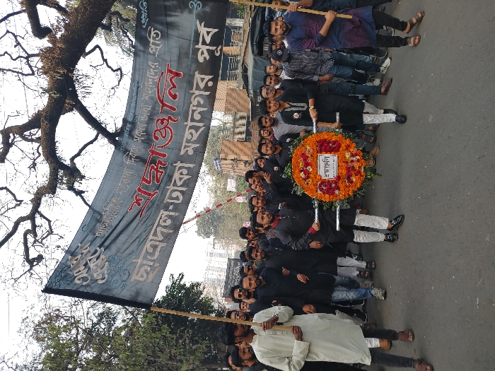 A group of people in Dhaka, Bangladesh, are marching with a banner written in Bengali. The banner appears to be for the student wing of a political party, "Chhatra Dal", and commemorates an event or leader. They are carrying a large, circular floral wreath with a central inscription in Bengali. The march is taking place outdoors on a street during daytime. Trees are visible on the sides, suggesting an urban setting.