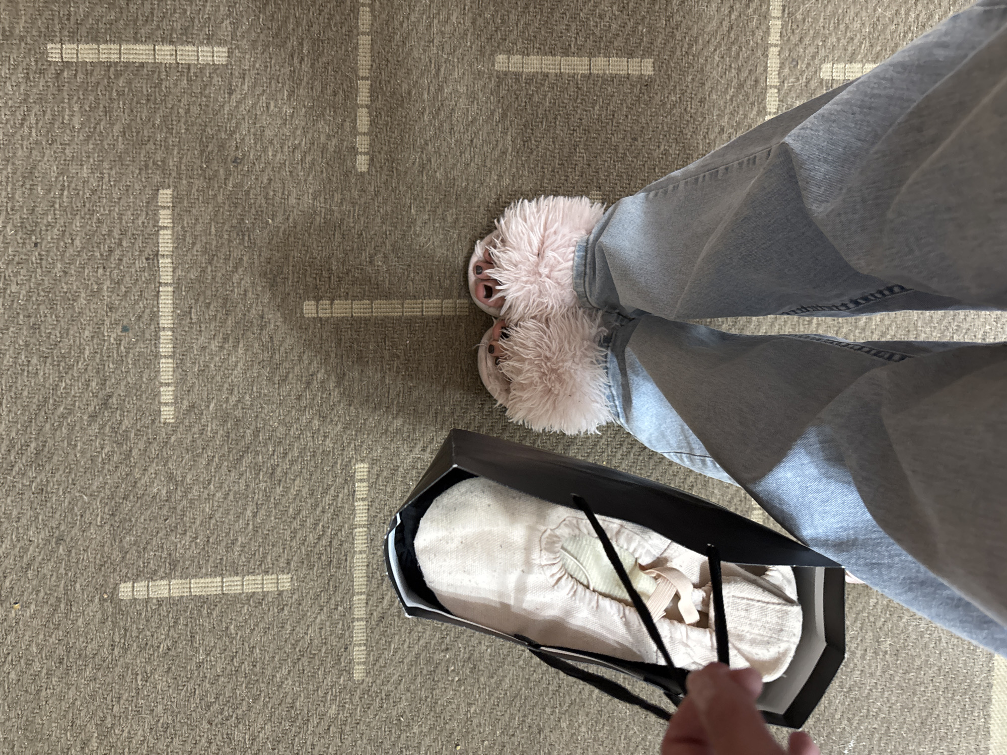 The image shows a person's legs and feet wearing light blue jeans and pink fluffy slippers, standing on a textured, grey carpet with a subtle grid pattern. To the left of the person's feet, a black box containing a pair of pink ballet shoes is visible. A hand is reaching into the box, holding onto a black ribbon. The scene appears to be indoors, likely in a residential setting in the city of Inskoy, Russia.