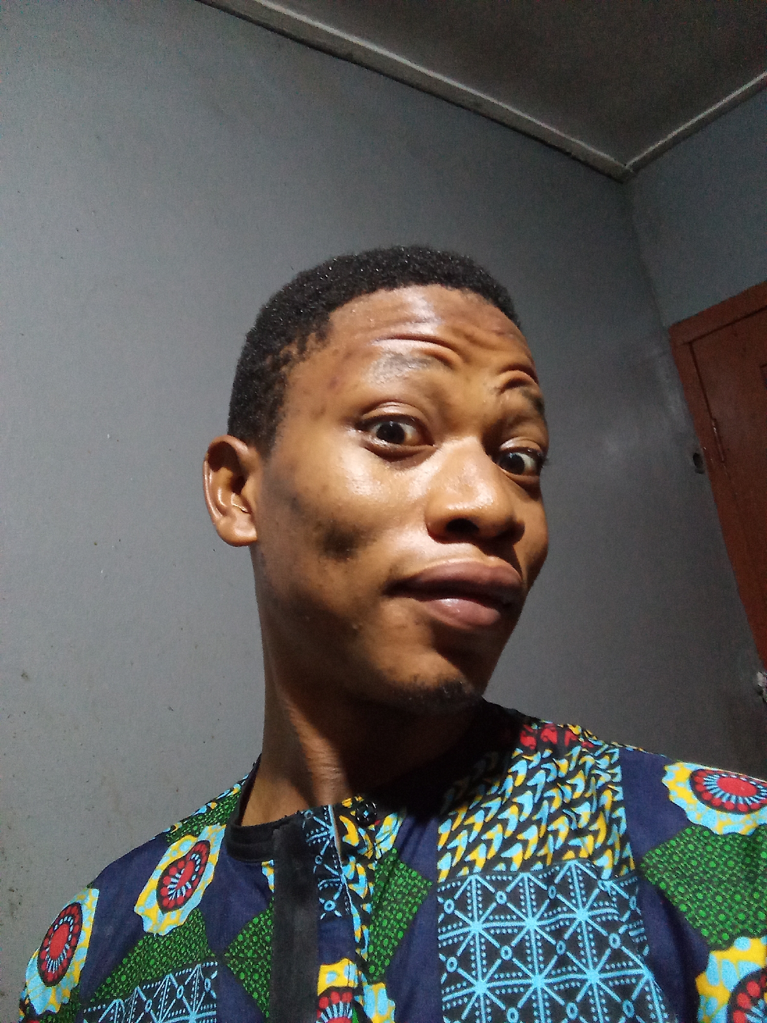 A medium indoor shot of a dark-skinned male with short black hair, wearing a blue, green, and yellow patterned shirt. The man is centered in the frame and appears to be taking a selfie. He has a slightly puckered expression and is looking directly at the camera with his eyes wide. The background consists of grey walls and a white ceiling molding. A brown door is visible in the upper right corner of the frame. The location is likely a residential interior but cannot be definitively determined to be in Oshogbo, Nigeria.