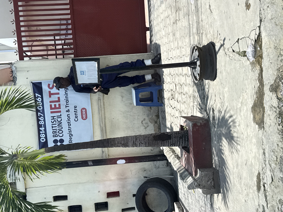 The image depicts an outdoor scene at the entrance of a facility identified by a banner as a "BRITISH COUNCIL IELTS Registration & Training Centre" in Alaguntan, Nigeria.
In the foreground, a concrete surface shows cracks and small puddles. A slender palm tree grows from a rectangular red concrete planter box on the left. To its left, a black vehicle tire rests against a light-colored building wall. To the right of the palm tree, a small, blue plastic stool is positioned on a paved area composed of interlocking blocks.
A black-framed banner is affixed to the building wall, displaying the phone number "0814-867-0367" and the text "BRITISH COUNCIL IELTS Registration & Training Centre," with a red box containing "HERE."
An adult male stands leaning against the wall, to the right of the banner and to the left of a large red metal gate that occupies the right side of the frame. He has dark skin, wears a dark baseball cap, a dark blue short-sleeved shirt, dark blue cargo pants, and dark shoes with white material visible above the ankles. A dark bag is slung over his left shoulder. He holds a small, dark object, possibly a mobile phone, in his left hand and gazes towards the left of the frame.
In the middle foreground, a dark metal pole with a rectangular sign attached stands anchored by a metallic wheel rim. The sign displays "CCII" at the top, with additional obscured text below. Shadows from the palm tree and the sign stand are cast on the ground, indicating bright overhead sunlight. A piece of white litter is visible on the cracked concrete near the sign's base.