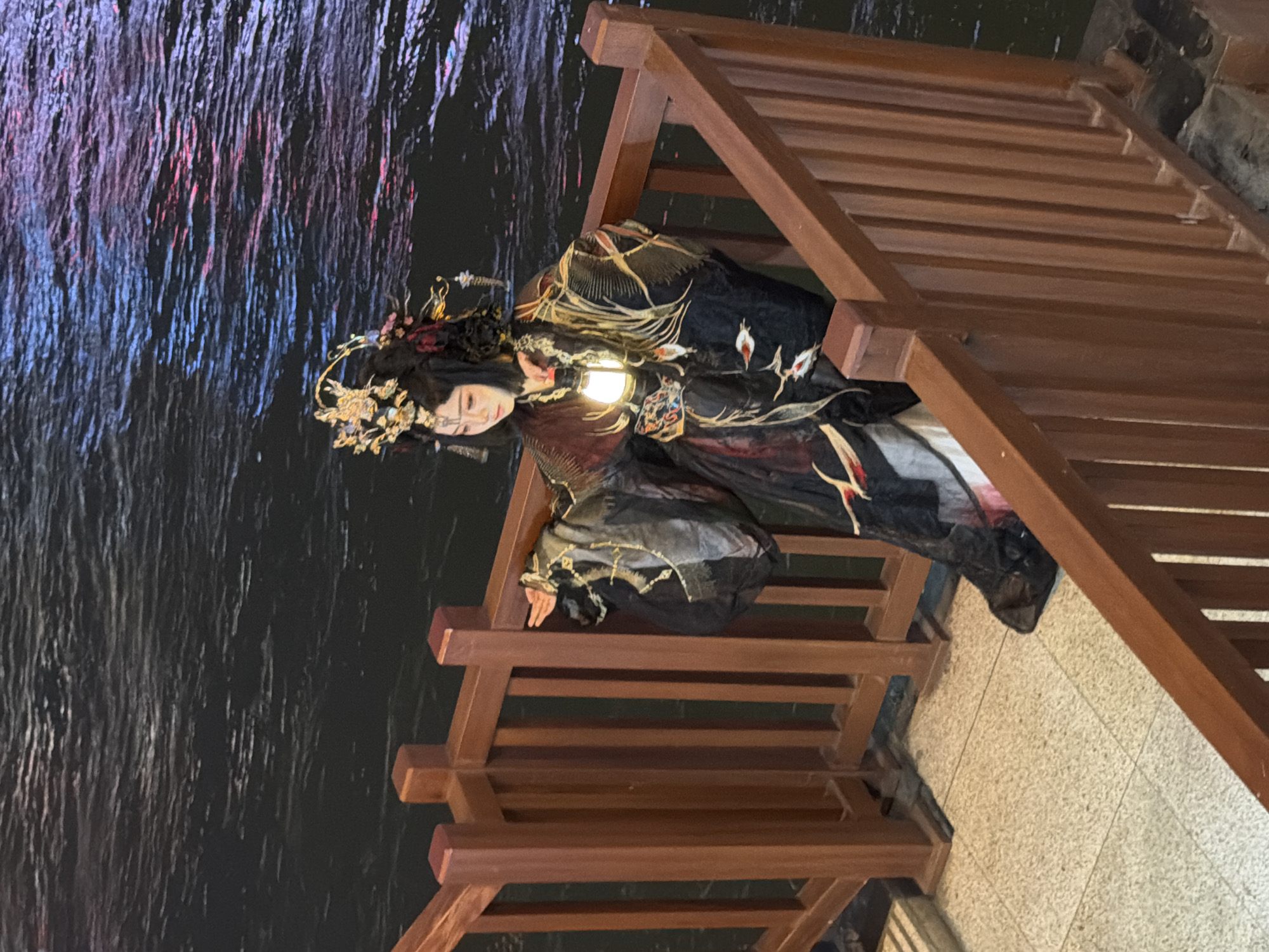 A person in traditional Chinese attire, possibly performing, sits on a tiled surface next to a wooden railing. The individual wears an elaborate headdress and dark, ornate clothing with gold and red accents. They are holding a glowing lantern in their left hand, and their right hand is extended outwards. The background features dark water with reflections of pink and blue lights, suggesting a nighttime scene. The location is identified as Yuzhong District, China.