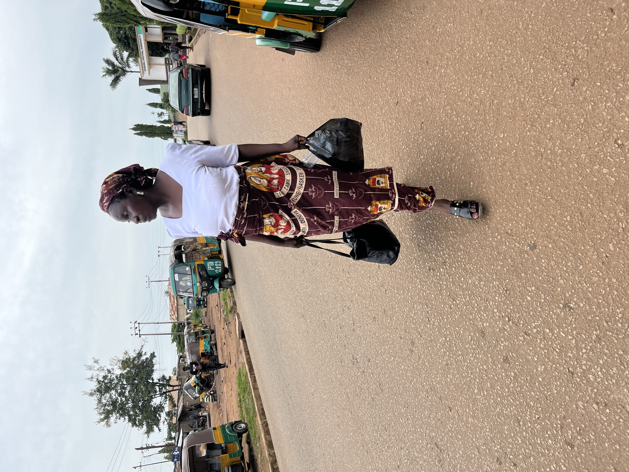 In Bukuru, Nigeria, a woman walks on a paved road, carrying two black bags. She is wearing a white shirt and a patterned maroon skirt, with a matching headscarf. The scene is bustling with activity, featuring several auto-rickshaws, also known as Keke Napeps, parked and in motion. A green sedan is visible on the road behind her, and a building with the name "KUGEL HOTEL RESORT" is in the background. Power lines crisscross the sky above the various structures and vegetation.