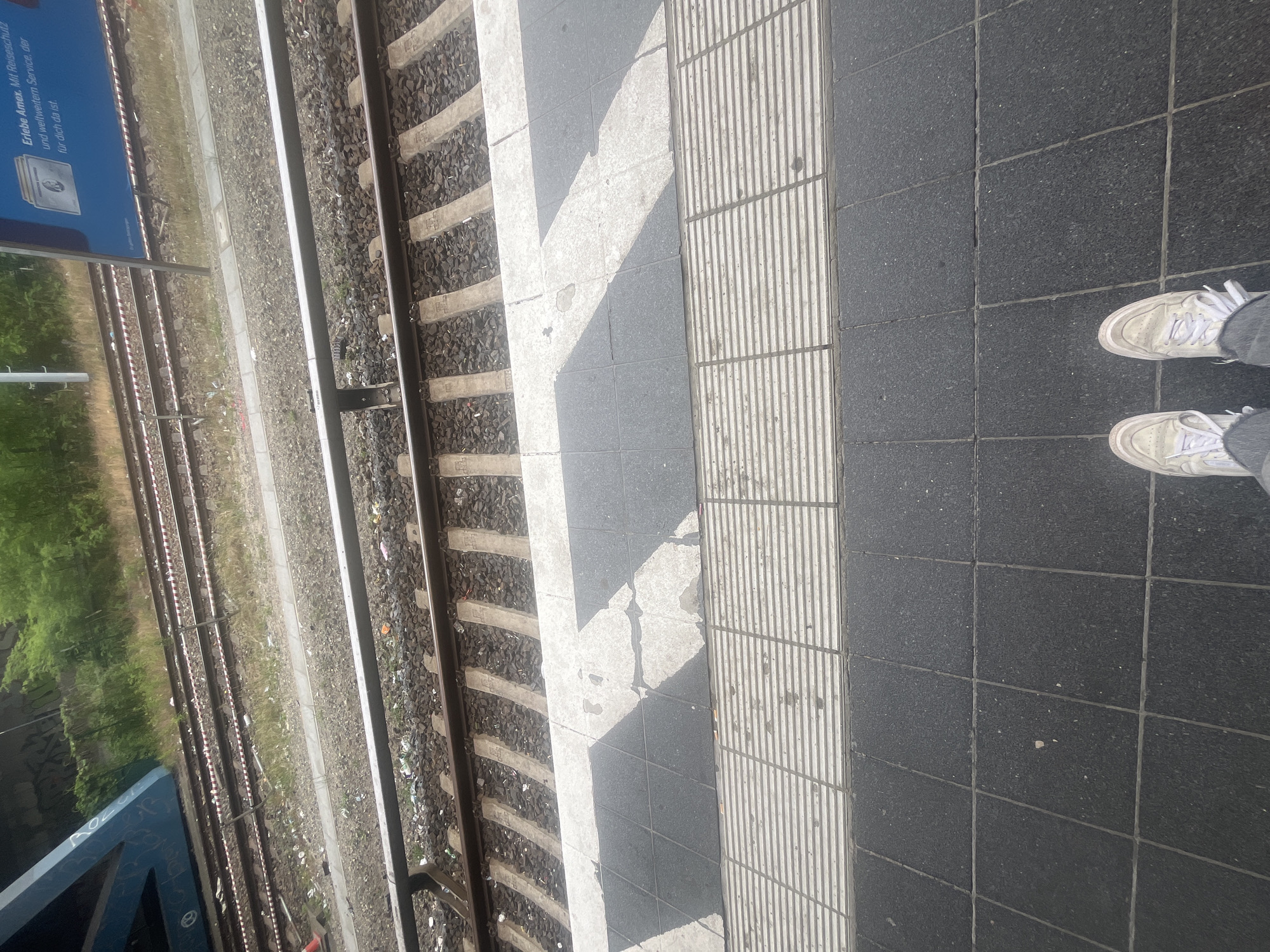 This image depicts a ground-level view of a train station platform in Berlin, Germany, looking down at the train tracks and the edge of the platform. In the foreground, a person's feet clad in white sneakers and gray pants are visible, standing on a dark tiled surface. The platform edge is marked by a textured, raised strip, followed by a section of dark and light patterned tiles. Beyond the platform, train tracks run parallel, with gravel ballast and concrete sleepers. Some debris, including plastic wrappers and cigarette butts, is scattered along the tracks and platform edge. In the background, a blue train carriage is partially visible, along with overgrown green vegetation and a section of a blue overhead structure with graffiti. A blue advertisement poster with German text is also visible on a pole to the right of the tracks. The overall scene suggests an urban outdoor environment at a public transportation hub.