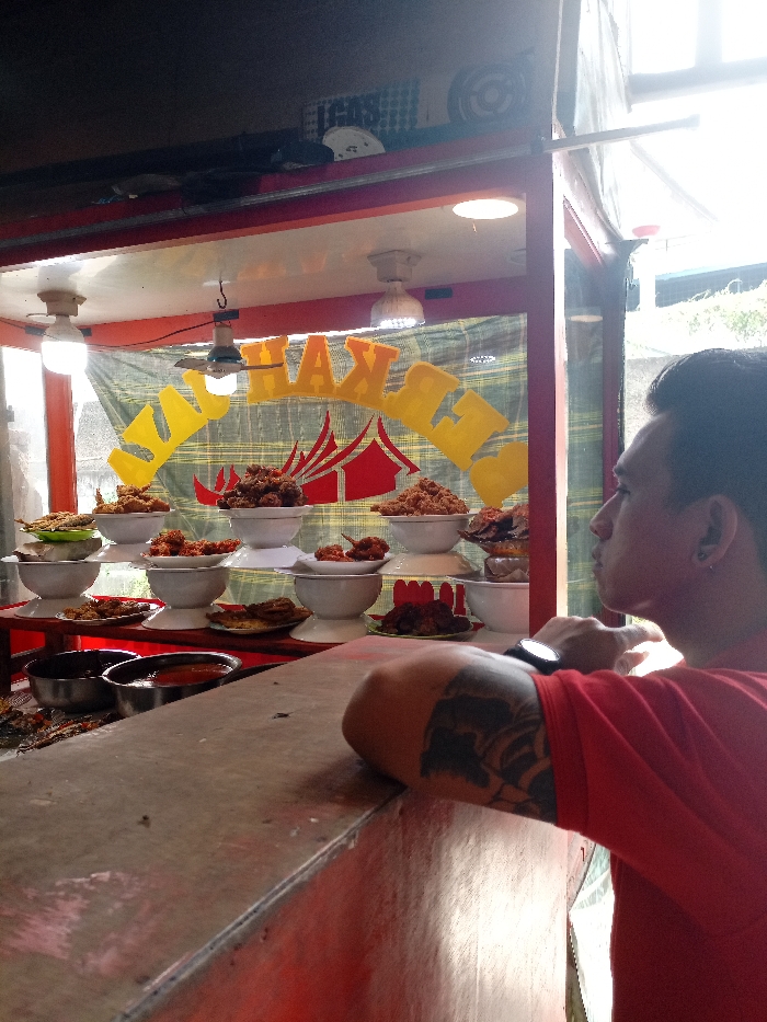 The image depicts a food stall, likely a "ayam penyet" stall, located at the coordinates -6.987, 107.553 in Katapang, Indonesia. The stall is made of red metal and features a yellow sign with the words "Ayam Penyet" written in yellow letters. It is partially enclosed with glass windows, with a brown wooden countertop. The stall is stacked with platters of food, likely containing deep-fried chicken and other accompanying dishes. A single man, wearing a red t-shirt, a black watch, and a tattoo on his left arm, is leaning on the countertop with his back to the camera, gazing at the food. His expression is contemplative. Behind him, a large window reveals a partially obstructed view of the street outside. The scene is lit by artificial lighting, casting a warm glow on the stall and the food.