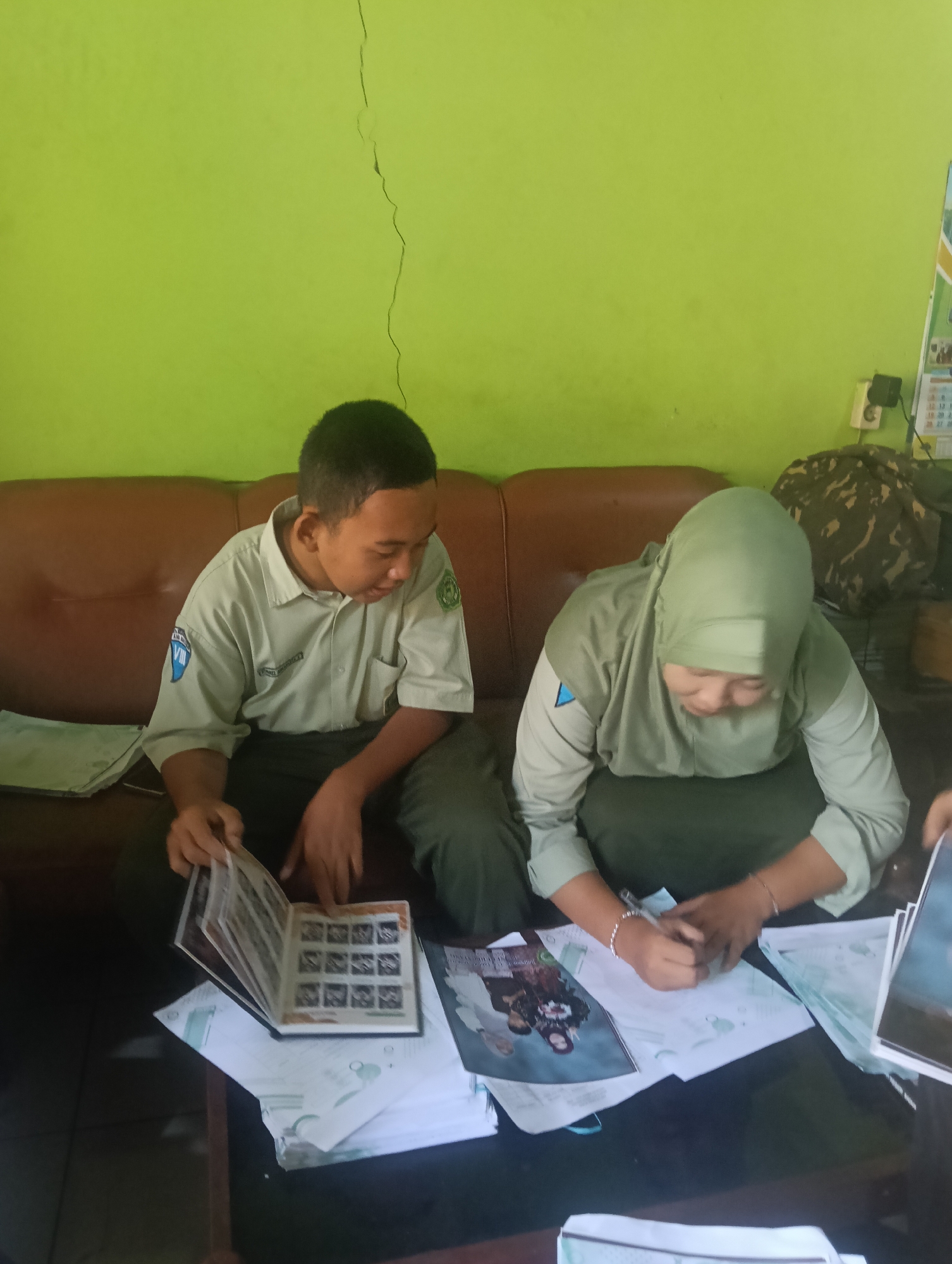 Two individuals, likely students based on their uniforms, are seated on a brown sofa in front of a lime green wall. The wall has a visible crack running vertically from the top. The individual on the left, wearing a light-colored shirt with emblems, is looking down at a photo album on a table in front of them. Their hands are near the album, and they appear to be interacting with its contents. The individual on the right, wearing a hijab and a similar uniform, is leaning forward and writing on a piece of paper on the same table. There are several papers and what appears to be another booklet or magazine on the table as well. A camouflage bag and a calendar are visible in the background on the right side of the image, suggesting an indoor setting. The overall scene suggests an educational or administrative task being performed.