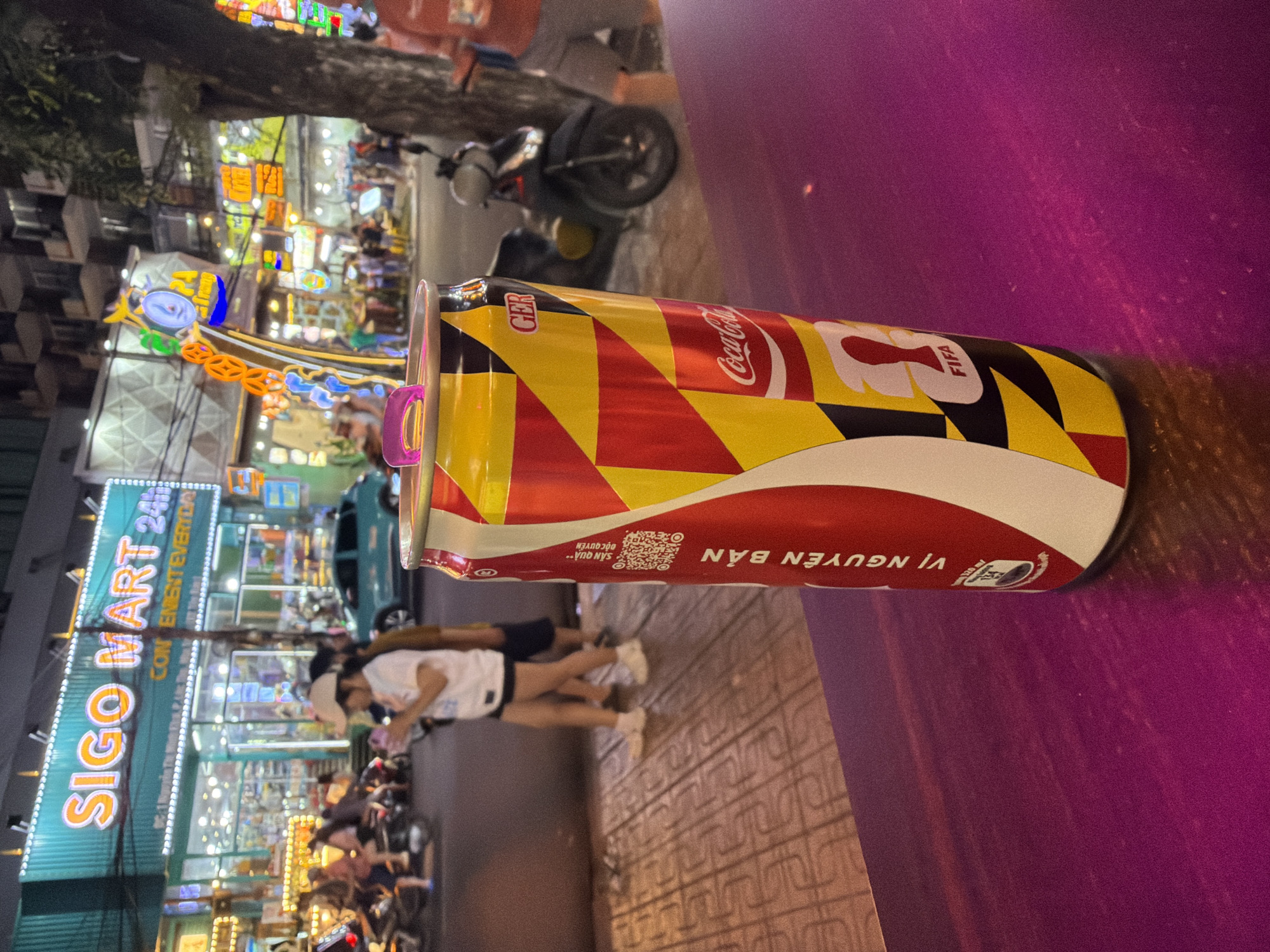 This media file is a close-up, sideways shot of a Coca-Cola can with a distinct, colorful design, positioned in the foreground. The can features a pattern of red, yellow, and black geometric shapes, along with the Coca-Cola logo and "FIFA" text, suggesting a promotional collaboration. Part of a purple surface occupies the right side of the frame, possibly a table.
In the background, a busy street scene unfolds at night. Neon lights illuminate storefronts and signs, creating a vibrant, urban atmosphere. A prominent sign reads "SIGO MART 24H CONVENIENT EVERYDAY," indicating a convenience store. Several motorcycles are parked along the street, and people can be seen walking and interacting in front of the store. The presence of motorcycles and the signage in English and what appears to be Vietnamese suggest a location in Vietnam. The overall impression is a lively evening in a commercial area, likely a city street. The lighting suggests it is nighttime, with artificial lights dominating the scene. No specific weather conditions are evident.