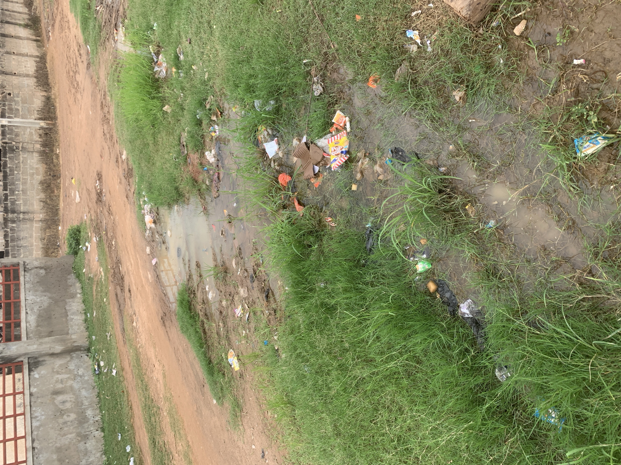 In Lafia, Nigeria, a muddy area with standing water is visible. The ground is uneven and appears to be a dirt path or roadside, littered with a significant amount of trash. Among the debris are plastic wrappers, paper packaging, and discarded food items. Lush green grass grows around the edges of the muddy patches. In the background, a rough concrete wall with a red-barred structure runs along the top. Further back, a brick building with a rough texture is visible. The scene suggests an area with poor waste management and potential drainage issues.