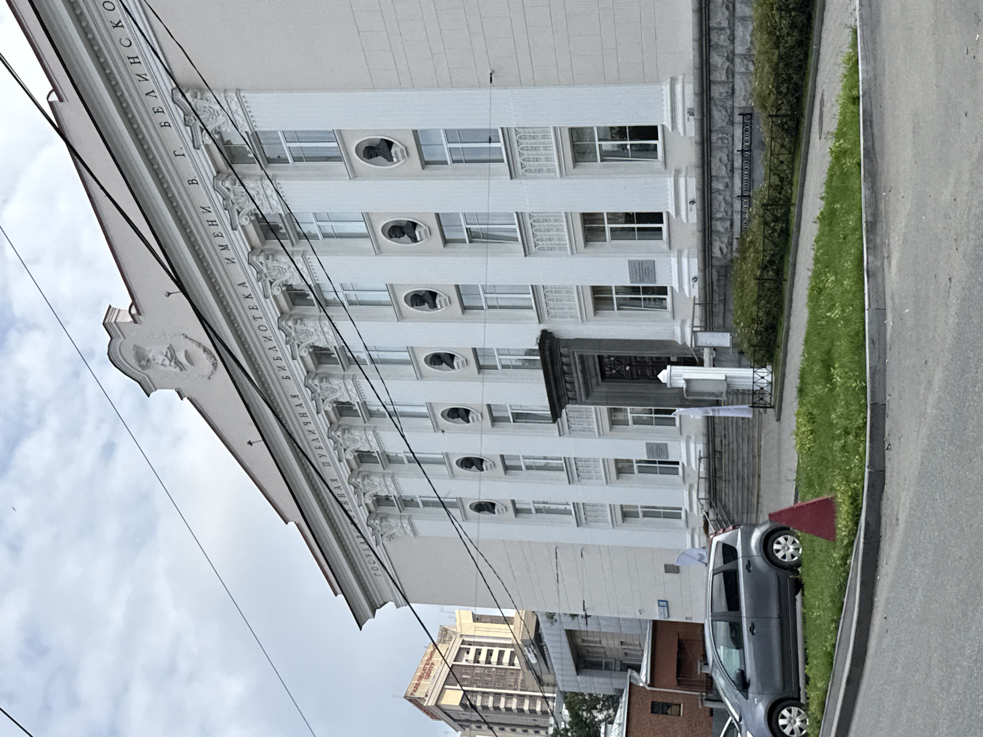 The image shows the facade of a large, classical-style building, identified by the inscription "ГОСУДАРСТВЕННАЯ ПУБЛИЧНАЯ БИБЛИОТЕКА ИМЕНИ В. Г. БЕЛИНСКОГО" (V.G. Belinsky State Public Library). The building is light-colored with prominent white columns and decorative architectural elements. Busts of notable figures are set into oval frames above the windows on the upper floor. The building is situated in Yekaterinburg, Russia. In the foreground, a gray minivan is parked on the side of a road. A red traffic cone is placed on a grassy area between the road and the building's entrance. Several power lines are visible crisscrossing the sky above the building. In the background, other urban buildings are visible, including a taller, modern yellow building. The sky is overcast.