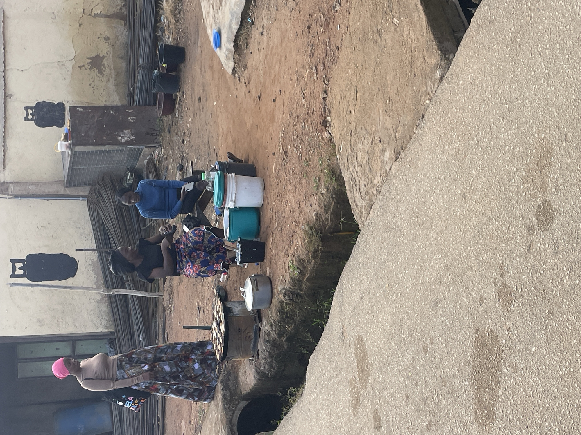 In Rafin Par, Nigeria, an outdoor scene unfolds with three women engaged in activities related to food preparation and vending. A woman on the left, wearing a pink headscarf and a long, patterned skirt, stands near a cooking stove laden with small, round food items. Beside her, a black bag with colorful lettering is slung over her shoulder. Two other women are seated. One, dressed in a black top and a floral skirt, appears to be attending to something in front of her. The other, in a blue long-sleeved shirt and dark pants, is seated and holds what appears to be a mobile phone. The environment is characterized by a dirt ground, with stacks of metal rods and construction materials visible in the background. A concrete structure, possibly a building, forms the backdrop. Several buckets and pots are placed around the women, suggesting their use in the food vending operation. A drainage channel runs along the foreground, with a concrete walkway alongside it. The overall impression is a snapshot of daily commerce in a Nigerian urban setting.