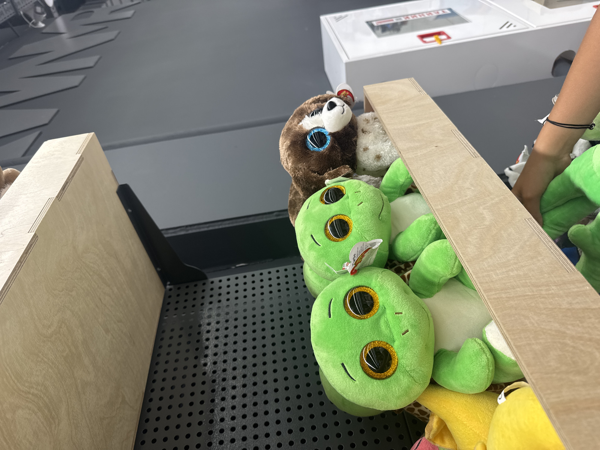 This media file shows a close-up view of a collection of plush toys, primarily green frog-like creatures and a brown sloth, neatly arranged in a wooden bin. The bin itself appears to be part of a retail display, possibly in a store or amusement center, given the black perforated surface below and a white electronic device with red and yellow accents visible in the background. A person's arm, adorned with a thin black bracelet, is reaching into the bin, suggesting they are selecting or arranging the toys. The lighting appears to be indoor, with no indication of the time of day or weather. The overall scene conveys a sense of playful consumerism or perhaps the organization of merchandise. The plush toys, with their large, glittery eyes, are the dominant objects, creating a bright and inviting visual. Some of the toys have small tags attached, typical of retail items. No discernible text is clearly legible within the frame, aside from what might be on the electronic device in the background, which is too indistinct to read.