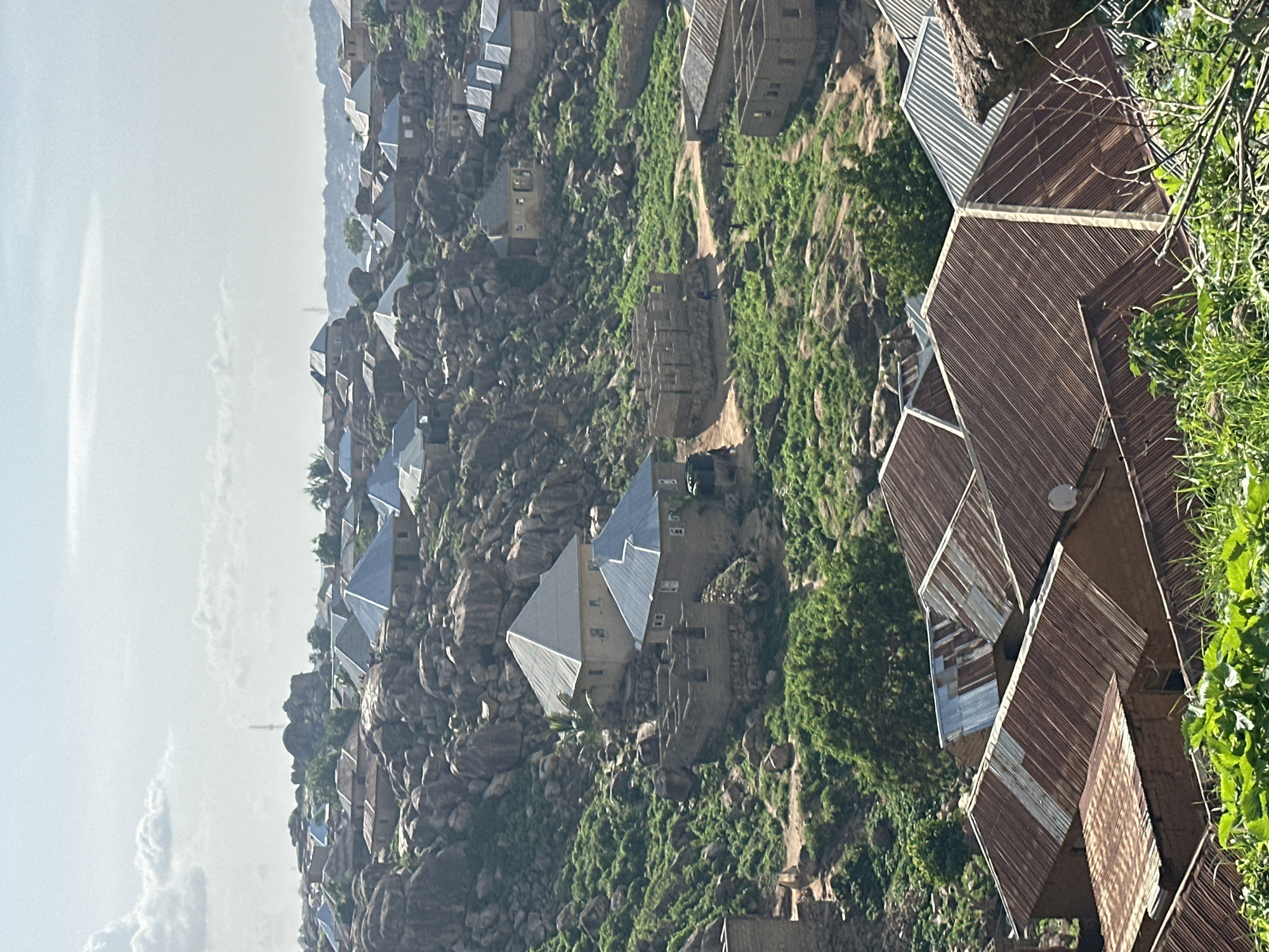 The image depicts a view of a residential area nestled on a rocky, hilly terrain. The architecture consists primarily of low-rise buildings with corrugated metal roofs, many of which appear weathered and rusted. Some structures are under construction, with visible brickwork and incomplete walls. Green vegetation, including grass and trees, is scattered across the landscape, growing amongst the rocks and around the buildings. The sky is overcast with diffused sunlight. The overall impression is of a community built into a challenging, natural environment. The style of construction and the landscape are consistent with the rocky hills and settlements found in and around Jos, Nigeria.