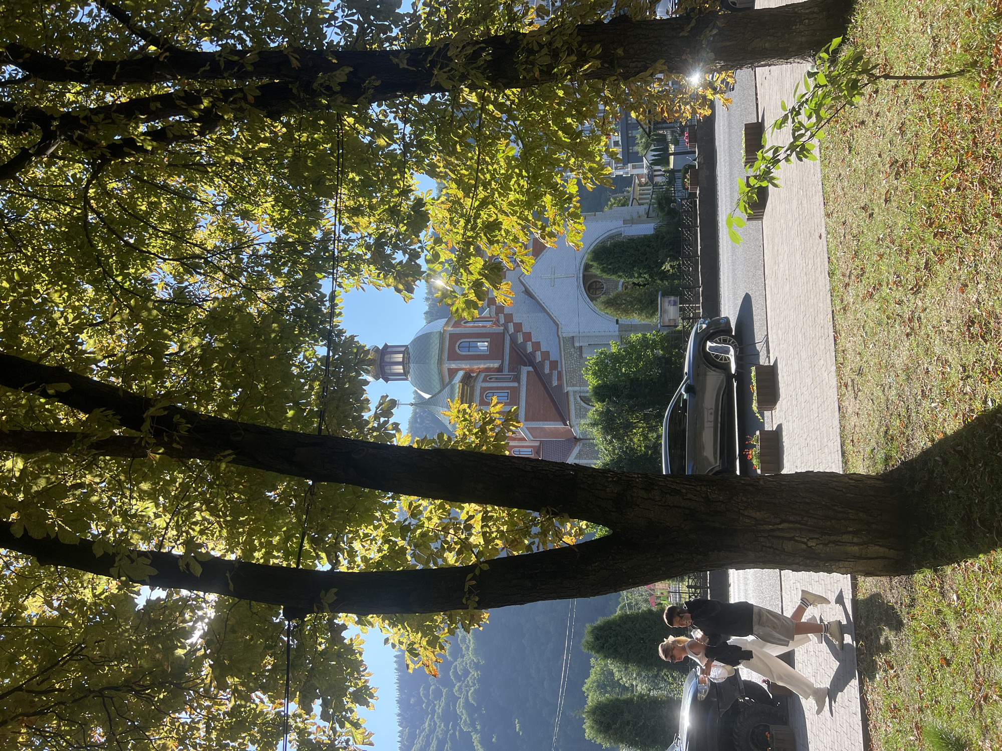 The image shows a sunlit scene in the city of Yaremche, Ukraine, likely in autumn given the yellowing leaves on the trees. In the foreground, two young people are walking along a paved pathway. The person on the left is wearing white pants and a white top, with blonde hair pulled back. They are carrying a bag in their left hand. The person on the right is wearing shorts and a dark hooded jacket, and appears to be holding a phone to their ear. They are walking from left to right in the frame. Behind them, a dark-colored SUV is parked on the left, and a darker sedan is driving or parked on the road to the right. The pathway is bordered by a grassy area with fallen leaves.
In the background, a large, ornate church with a green dome and red and white facade is visible, partially obscured by the trees. The church has a cross on its highest dome. Beyond the church, a densely forested hillside rises under a clear blue sky. The sun is shining brightly, casting shadows from the trees onto the pathway and ground. The overall impression is a pleasant, possibly late afternoon or early morning, scene in a town with significant natural beauty and a prominent religious structure.