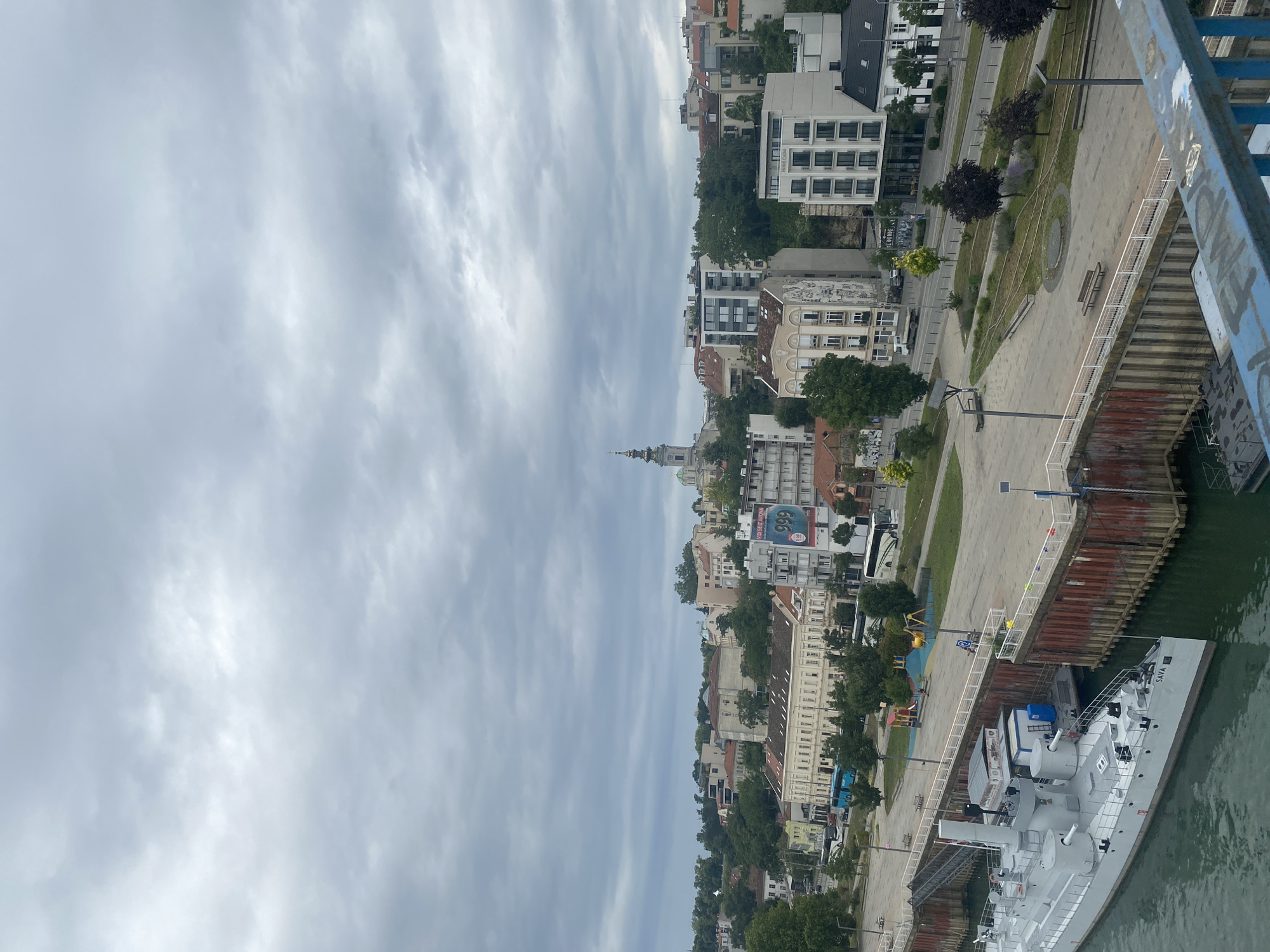 The image captures a cloudy day overlooking the Sava River in Belgrade, Serbia. In the foreground, the bow of a gray ship labeled "SAVA" is docked. Behind the ship, a wide promenade area with paved surfaces and green spaces is visible. A playground with colorful equipment is situated on the promenade.
Beyond the promenade, a city skyline rises, characterized by a mix of architectural styles. Numerous buildings, varying in height and color, are nestled amongst lush green trees. Some buildings exhibit classical European architecture with ornate facades, while others are modern high-rises. A prominent church steeple with a green dome can be seen in the distance, suggesting a religious or historical landmark. Various vehicles, including buses and cars, are visible on the road running parallel to the promenade. The sky is overcast with heavy gray clouds, creating a subdued and diffused light. The overall scene depicts a riverfront area in a European city, likely a historic district due to the blend of old and new architecture.