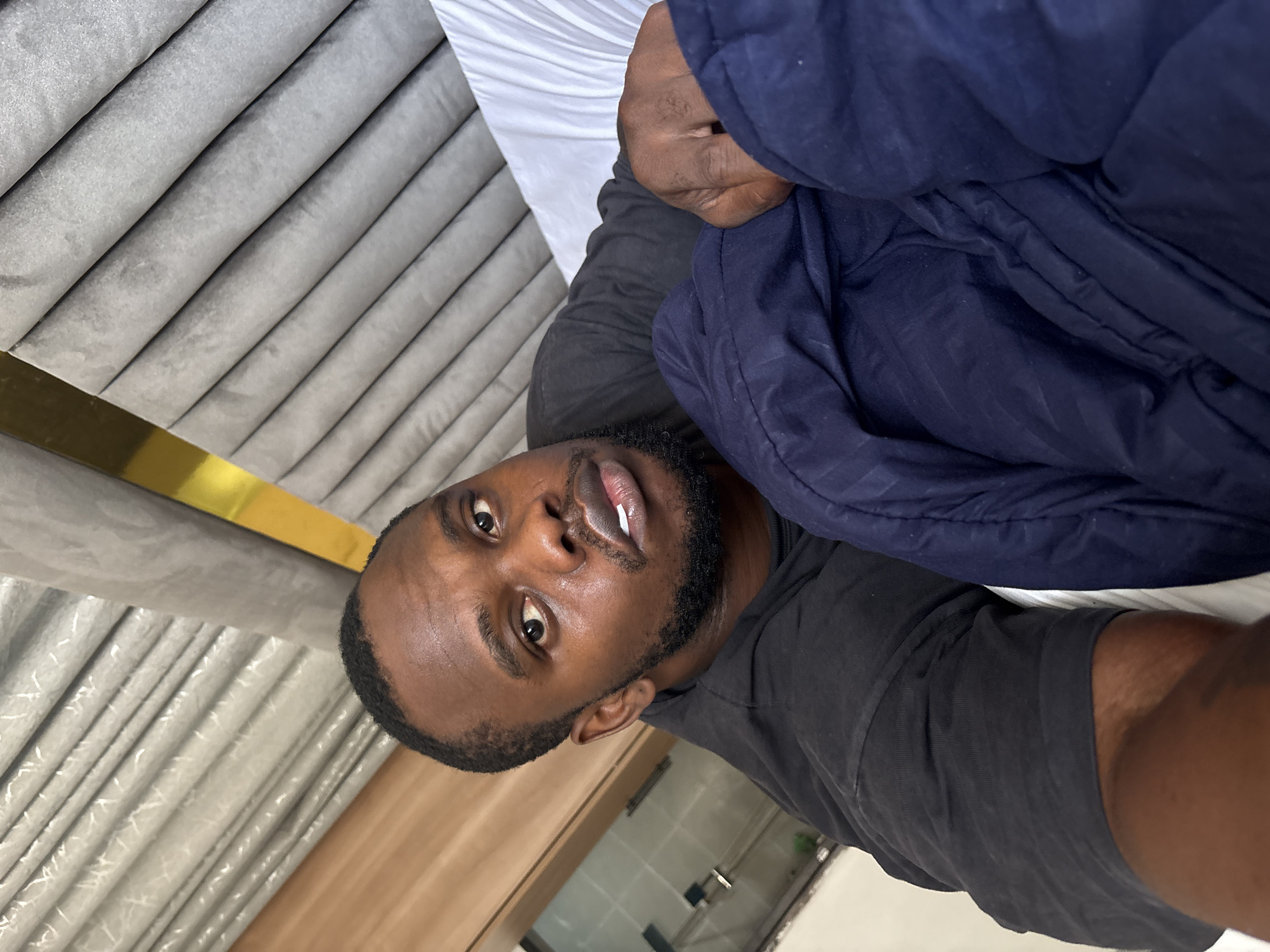 A man with dark skin, a beard, and a black t-shirt takes a selfie indoors. He is partially covered by a dark blue puffy jacket. The background includes a grey, upholstered headboard with gold trim, a white sheet, and a glimpse of a bright, possibly tiled, room. The location, Iddo Tudunwada, Nigeria, cannot be confirmed from visual information.