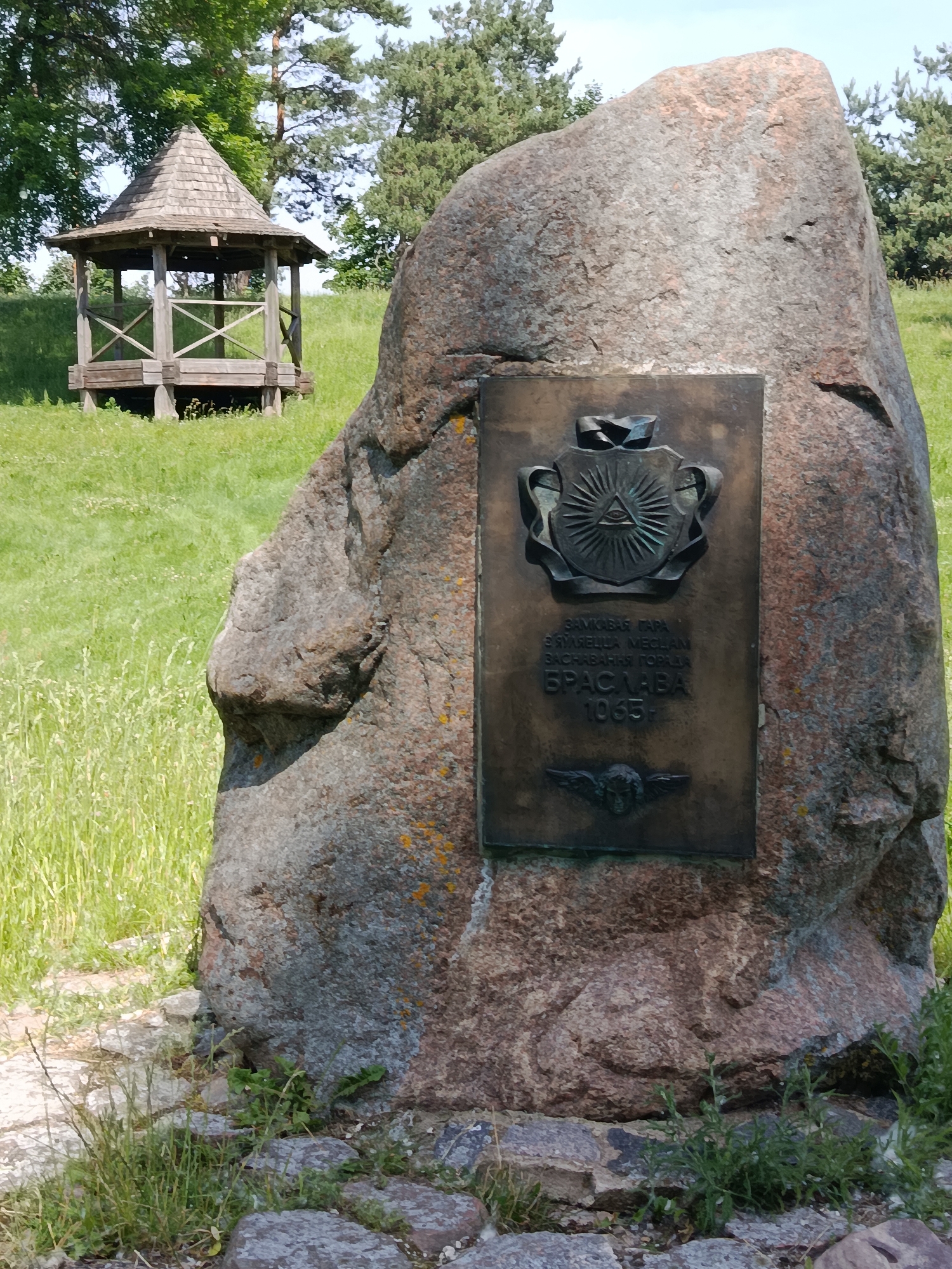 A granite boulder with a bronze plaque is situated in a grassy area. The plaque features a coat of arms with an all-seeing eye symbol at its center, and text below that reads "ЗАМКОВАЯ ГОРА / З'ЯЎЛЯЕЦЦА МЕСЦАМ / ЗАСНАВАННЯ ПОРЯДКА / БРАСЛАВА / 1065". A small winged human head is depicted at the bottom of the plaque. In the background, a wooden gazebo with a conical roof stands on a grassy hill. The scene is set outdoors during the day.