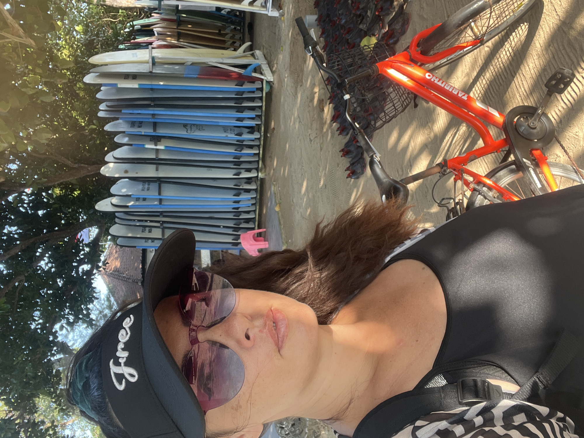 The image shows a woman in a black visor, sunglasses, and a black tank top, taking a selfie. Behind her, a rack of surfboards stands on a sandy beach. A red bicycle with a basket is parked in the foreground. In the background, there is a small pink stool and trees. The scene is lit by sunlight, casting shadows on the ground. The presence of surfboards and the sandy beach suggests a coastal location, likely in Kuta Selatan, Indonesia, a popular tourist destination known for its beaches and surf spots.