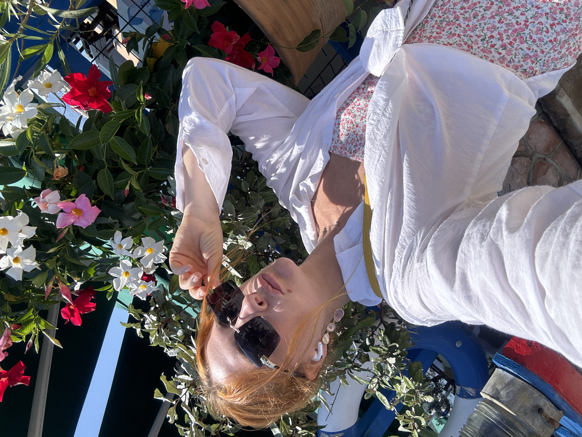 The image shows a woman with red hair wearing sunglasses, a white unbuttoned shirt, and a floral-patterned dress. She is holding her sunglasses with her left hand and has white wireless earbuds in her ears. She is leaning against a railing that has a blue and red structure attached to it, and a plant with red, white, and pink flowers is growing around her. The background appears to be outdoors, with sunlight casting shadows. The setting resembles a public space with greenery and some industrial-looking elements. Given the presence of a yacht club structure in the background and the overall aesthetic, this scene is likely captured at the Budva Yacht Club in Budva, Montenegro, a popular coastal city known for its scenic beauty and vibrant atmosphere.