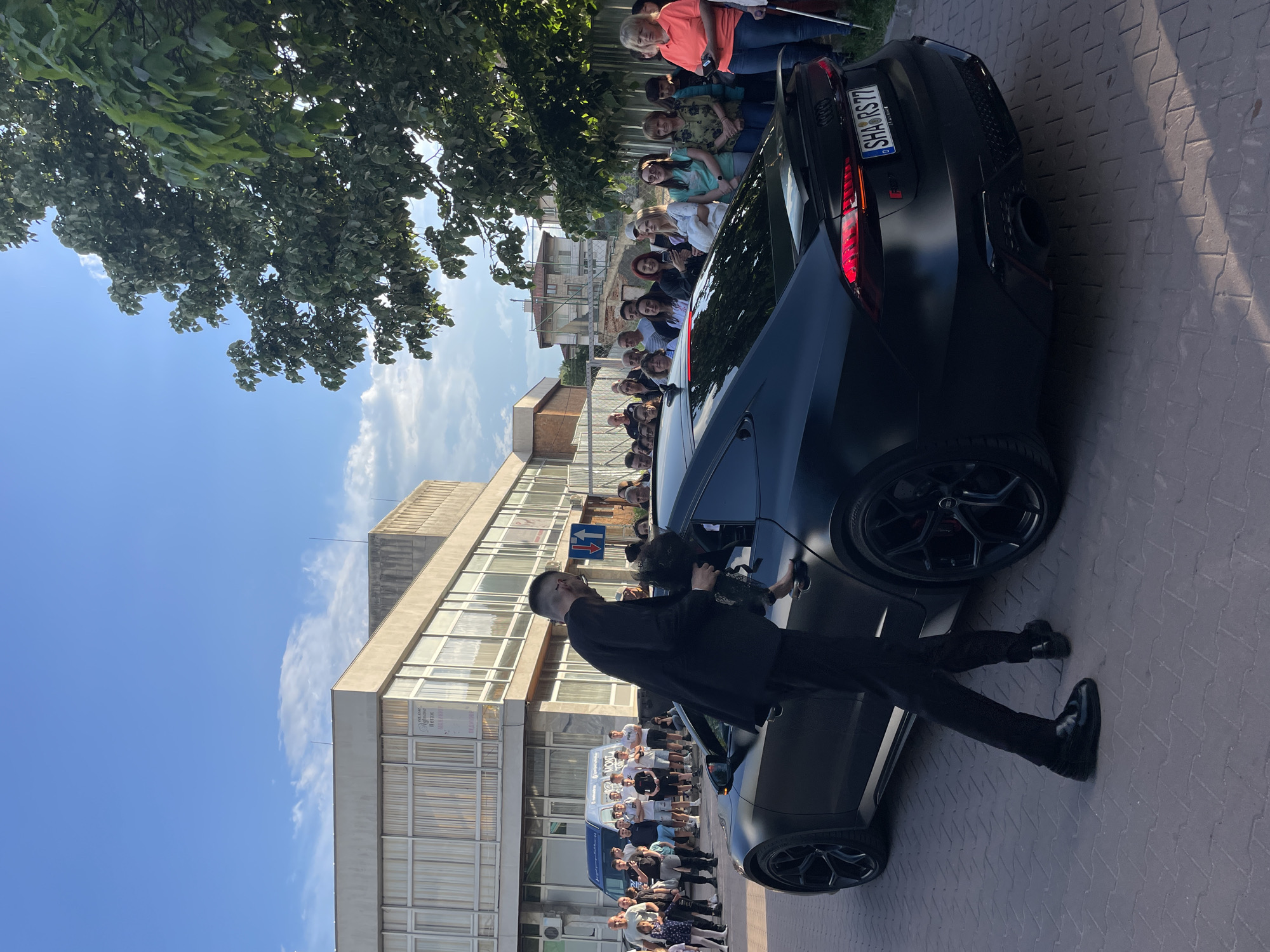 The image shows a black car, possibly an Audi RS, parked on a brick-paved area in Zlatograd, Bulgaria. People are gathered around the car, some of whom are taking pictures. A man in black clothing stands near the car. In the background, there is a building with a multi-story window facade. The license plate reads "SHA RS 77". The scene is outdoors during daylight.