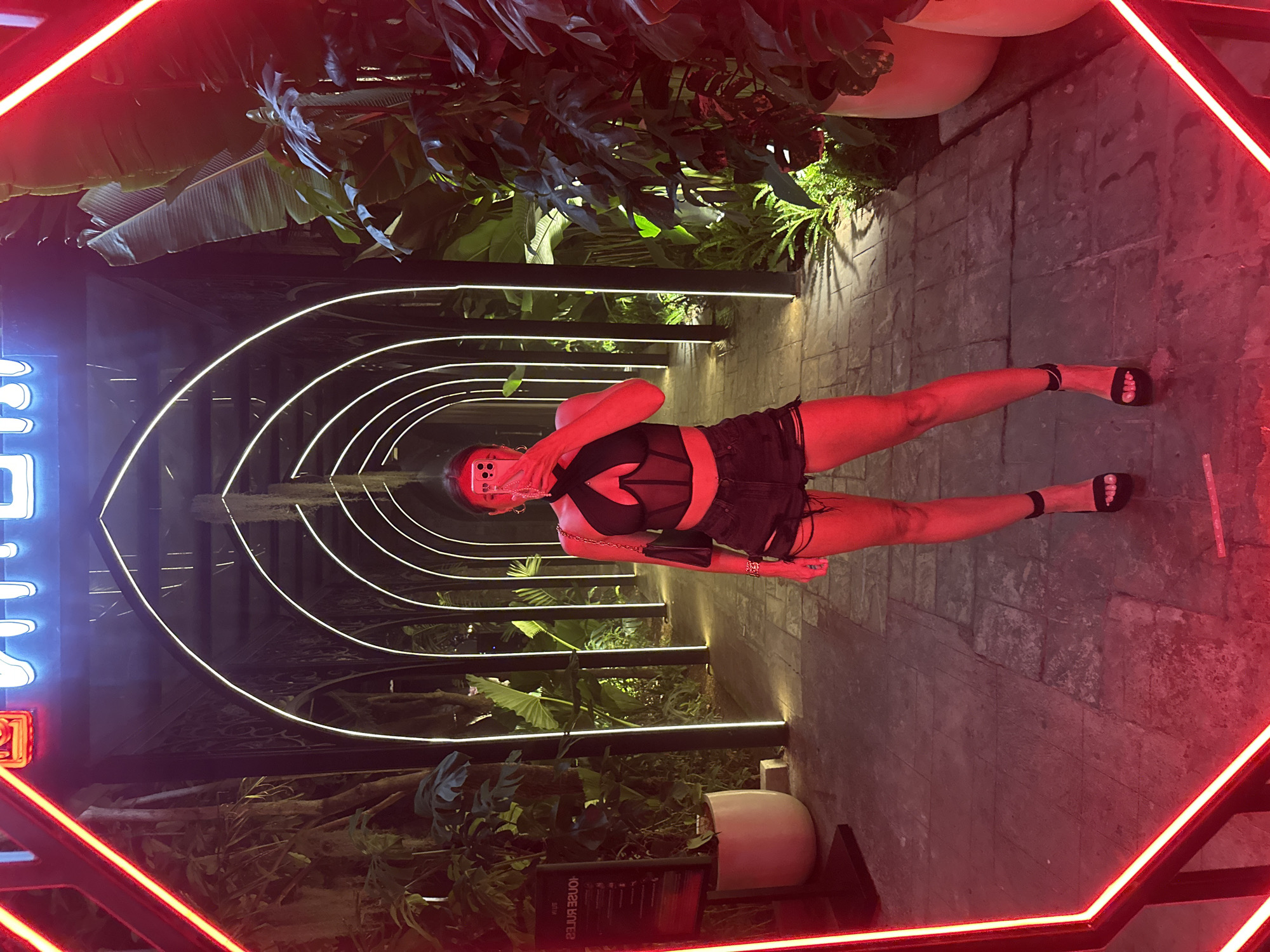 A woman is taking a selfie in a mirrored hallway with neon red lighting and tropical plants. She is wearing a black corset-style top, ripped black shorts, and black platform heels. The hallway has arched openings with white lights. There are large potted plants and trees on either side of the walkway. A sign with text is visible on the left side of the image. The scene appears to be in a nightlife establishment in North Kuta, Indonesia, suggested by the combination of lush vegetation, modern lighting, and a potentially upscale ambiance.