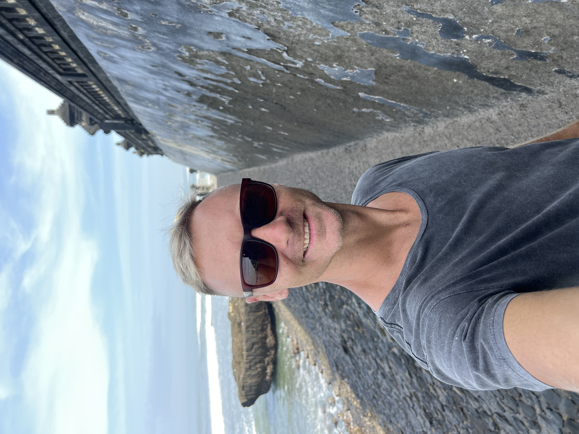 A man with short, blonde hair and sunglasses takes a selfie on a cobblestone path with the ocean on his left and a textured, weathered wall on his right. The wall appears to be part of a large, dark, ornate structure, possibly a temple or historical building, with decorative elements visible at the top. The sky is overcast with a few streaks of blue. In the background, there's a large rock formation in the ocean, and waves are breaking on the shore. The man is wearing a dark grey t-shirt. The location is likely the Tanah Lot temple complex in North Kuta, Bali, Indonesia, known for its coastal setting and distinctive architecture.