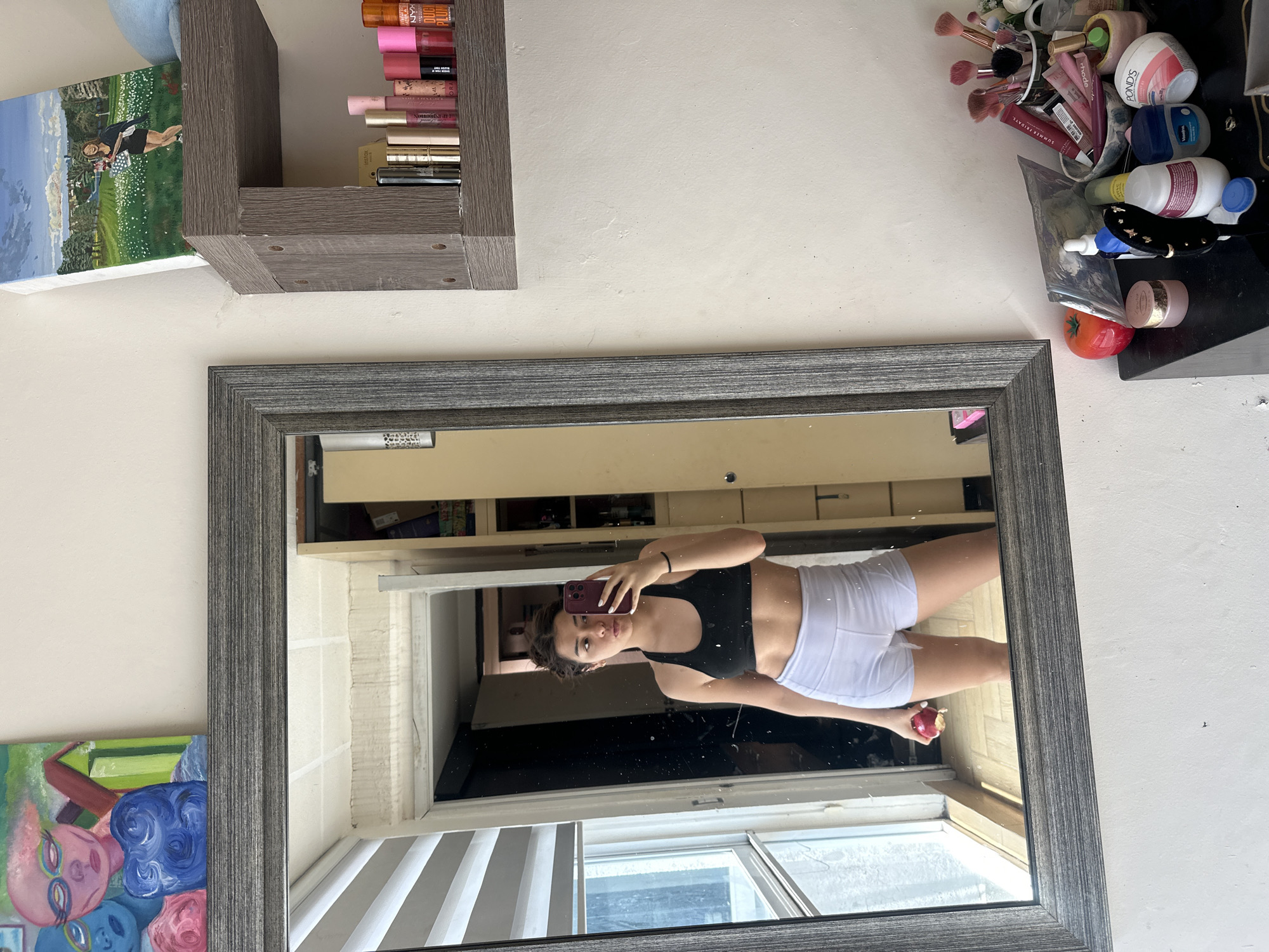 A reflection in a grey-framed mirror captures a young woman in workout attire—a black sports bra and white shorts—holding a cell phone in one hand and a red apple in the other. The mirror hangs on a white wall, positioned between a shelf filled with makeup items and a painting of whimsical figures. The reflection includes a glimpse of a bright, window-lit space with blinds, suggesting an interior. The reflected scene hints at a city location through the window, but there is no additional evidence indicating the location is Puebla, Mexico.