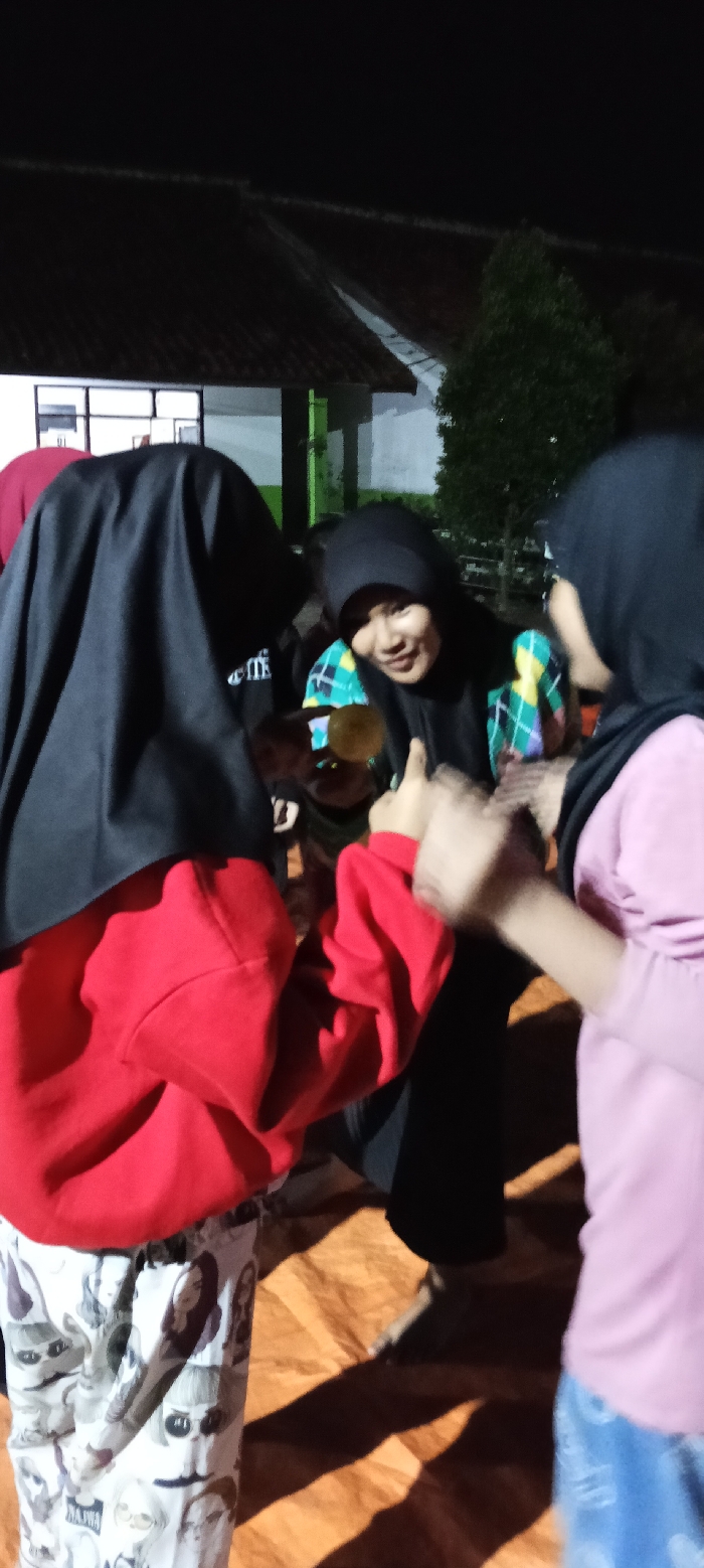 The image shows three young women standing in a dimly lit outdoor setting, possibly at night. The women are all wearing headscarves. Two of the women are interacting, with one of the women, wearing a red sweatshirt and patterned pants, smiling and looking at the other woman, wearing a light pink long-sleeved shirt and blue jeans, while holding her hand. The woman in pink is looking towards the other woman and smiling. The other woman is out of focus and is wearing a black hijab and a plaid shirt over it, smiling and looking down towards the other woman’s hand. The women are standing on a wooden surface, and a dark shadow is cast on the surface in front of them. There is a building with a white wall and a green-colored window to the left of the image. There is a small green tree in the background, and the sky is black. The setting is in Cangkuang, Indonesia, based on the coordinates provided.