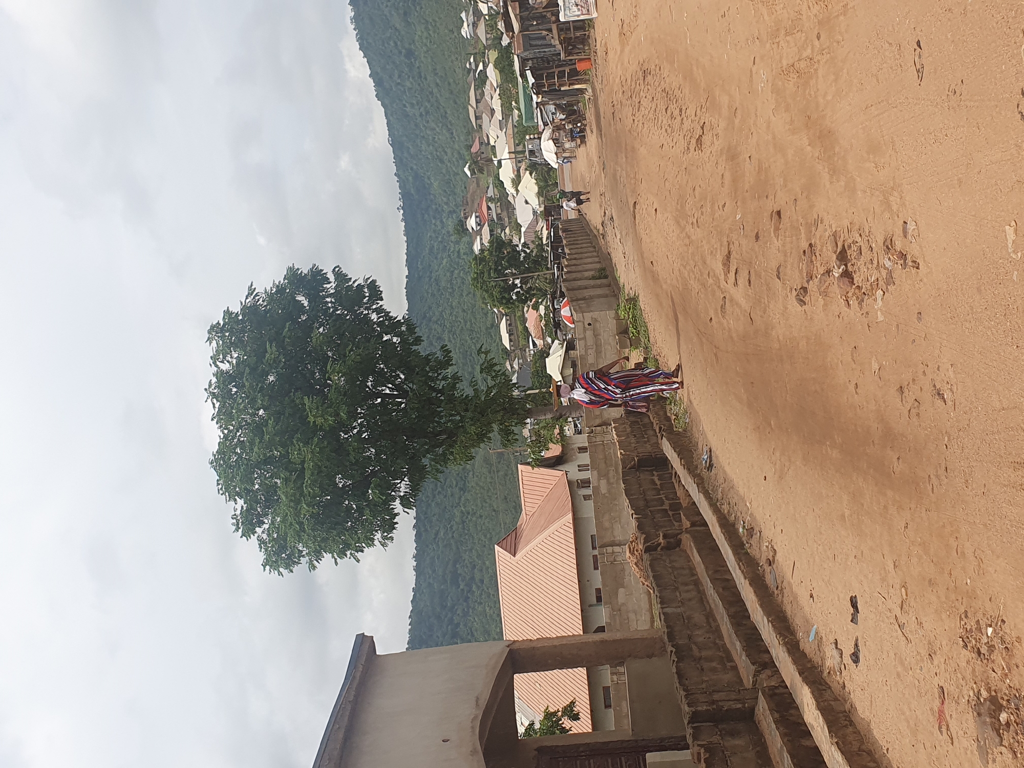 The image depicts a view in Lokoja, Nigeria, characterized by a dirt road leading towards a hillside densely populated with buildings. A large tree with green foliage is prominent on the left side of the frame. In the foreground, a dilapidated building structure is visible, with a person wearing a colorful striped cloth positioned near its edge. The overall environment suggests a rural or peri-urban setting within Lokoja.