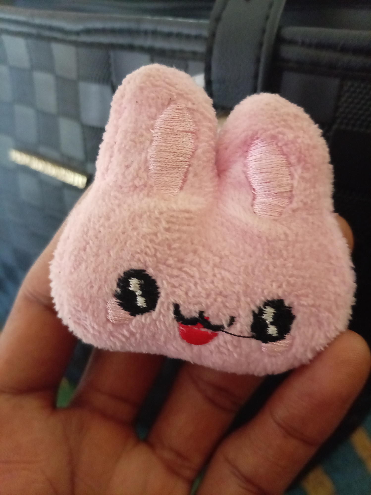 A close-up shot features a pink, plush bunny toy held in a human hand. The toy has embroidered features including black eyes, a red mouth, and pink ears. The background is partially visible, revealing a textured, dark-colored surface that appears to be part of a bag or other object. There is no indication of a location within Owerri, Nigeria, or any other place. No people or interactions are completely visible, just the single hand holding the toy.