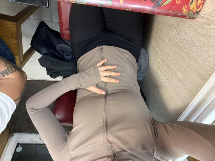 A person sits at a table, wearing black leggings and a tan zip-up jacket. A hand rests on the jacket. A bag lies to the left of the person, and a tattooed leg is visible. The table has a red patterned top. A wooden chair and white wall are nearby. The floor appears to be wooden.