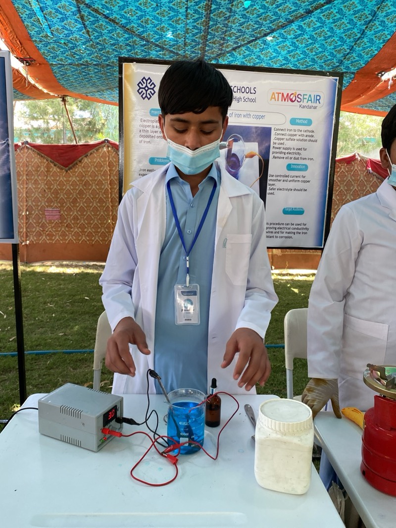 The image shows a young boy, likely a student, wearing a light blue shirt, a white lab coat, and a light blue surgical mask, standing behind a table outdoors. He has short dark hair and is looking down intently at a scientific experiment set up before him.
On the table, a gray power supply unit displaying "3.0" (presumably volts) is connected by red and black wires to a beaker filled with a bright blue liquid, indicative of a copper sulfate solution. Electrodes are submerged in the solution, suggesting an electroplating demonstration. To the right of the beaker, a small brown dropper bottle and a larger white plastic container are present. Another individual, also in a white lab coat, is partially visible on the right.
Behind the boy, a large informational poster is displayed, indicating the event is likely a science fair or exhibition. The poster is titled "ATMOSFAIR Kandahar" and features text related to a project on "Electroplating of iron with copper." Visible details on the poster include the method: "Connect iron to the cathode," "Connect copper with anode," "Copper sulfate solution should be used," "Power supply is used for providing electricity," and "Remove oil or dust from iron." It also lists "Innovation" points such as "Use controlled current for smoother and uniform copper layer" and "Safer electrolyte should be used." The "Target Audience" section states the procedure can be used for "proving electrical conductivity wires and for making the iron resistant to corrosion."
The scene is set outdoors under a patterned blue and orange tent or canopy, with green grass visible in the background, suggesting a clear, sunny day. The boy's posture and downward gaze indicate focus on the experiment, showcasing an educational activity in Kandahar, Afghanistan.