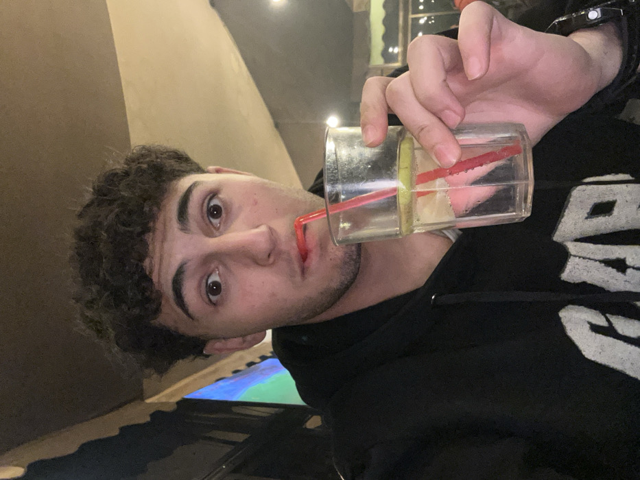 A young man with curly brown hair takes a sip from a clear glass using a red straw. The glass also contains a lemon wedge. He wears a black hoodie with white lettering. A black watch is visible on his wrist. The background is dimly lit, suggesting an indoor setting. The image is slightly tilted, with the man's face and the glass prominently featured.