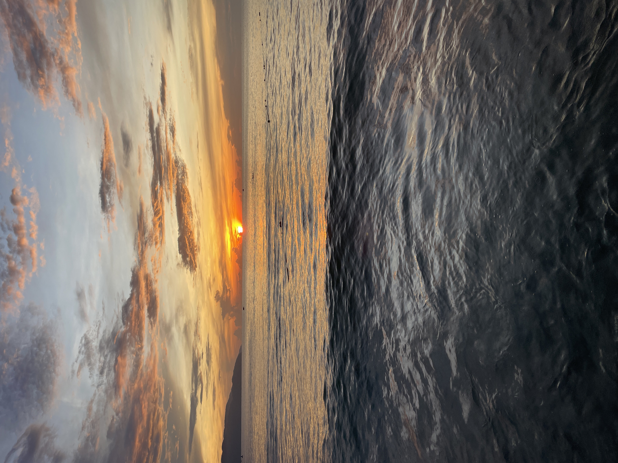 This is a vertical, full-frame photograph of a sunset over the ocean in Da Nang, Vietnam. The sun, a bright orange orb, is setting on the horizon, casting a warm glow across the sky and water. The sky is filled with clouds painted in hues of orange, pink, and yellow, with hints of blue at the top. The ocean is calm, with gentle waves reflecting the colors of the sunset. In the distance, a silhouetted mountain range forms the backdrop. A few buoys or markers are visible in the water, marking a boundary or pathway. The foreground of the image is dominated by dark, rippling water, creating a stark contrast with the vibrant sky. The scene evokes a sense of tranquility and natural beauty.