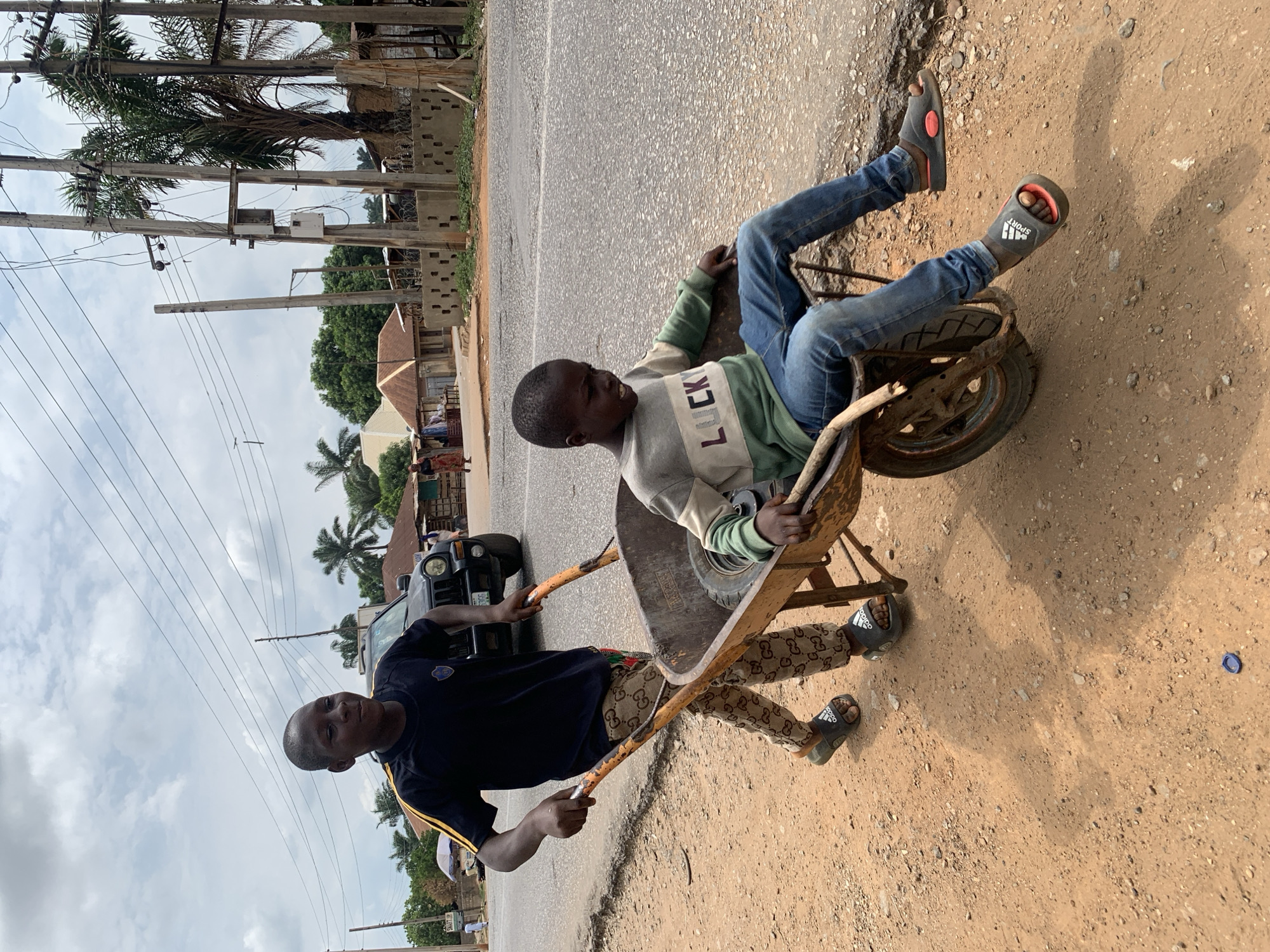 In Akwanga, Nigeria, two boys are on a road next to a dirt shoulder. One boy is sitting in an old, rusty wheelbarrow, wearing a "LUCKY" sweater, blue jeans, and Adidas sandals. The other boy is pulling the wheelbarrow, wearing a dark t-shirt with yellow stripes, patterned pants, and Adidas sandals. A black SUV is parked nearby, with power lines and palm trees visible in the background. The sky is cloudy.
