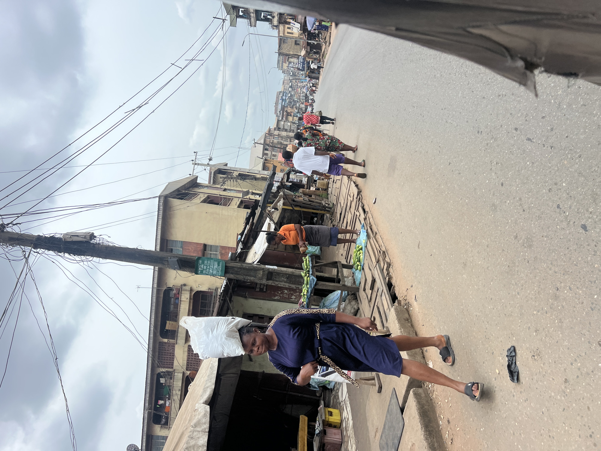 This media file captures a street scene in Nkpor, Nigeria. The primary subject is a woman in a dark blue dress with a leopard-print belt, carrying a large white bag balanced on her head. She is walking on a paved road, and her gaze is directed downwards. In the background, several other people are visible, some walking along the street and others appearing to be vendors.
The setting is a bustling urban environment, likely a market street. Buildings line both sides of the road, some appearing to be shops or residences, with signs and awnings indicating commercial activity. Several stalls are set up along the sidewalk, with one displaying what appears to be produce, possibly cucumbers. Electrical wires crisscross the sky, strung between poles, a common sight in many urban areas.
The weather appears overcast, with a muted sky suggesting it could be morning or late afternoon. The overall atmosphere suggests a typical day of commerce and movement in the area. A green sign with white text is visible on one of the buildings, but the text is largely illegible due to the image resolution and distance.
The image is captured from a slightly elevated perspective, possibly from a vehicle, as indicated by the partially visible dark frame at the top of the image. The rotation of the image suggests it was taken with a phone camera and then not reoriented.