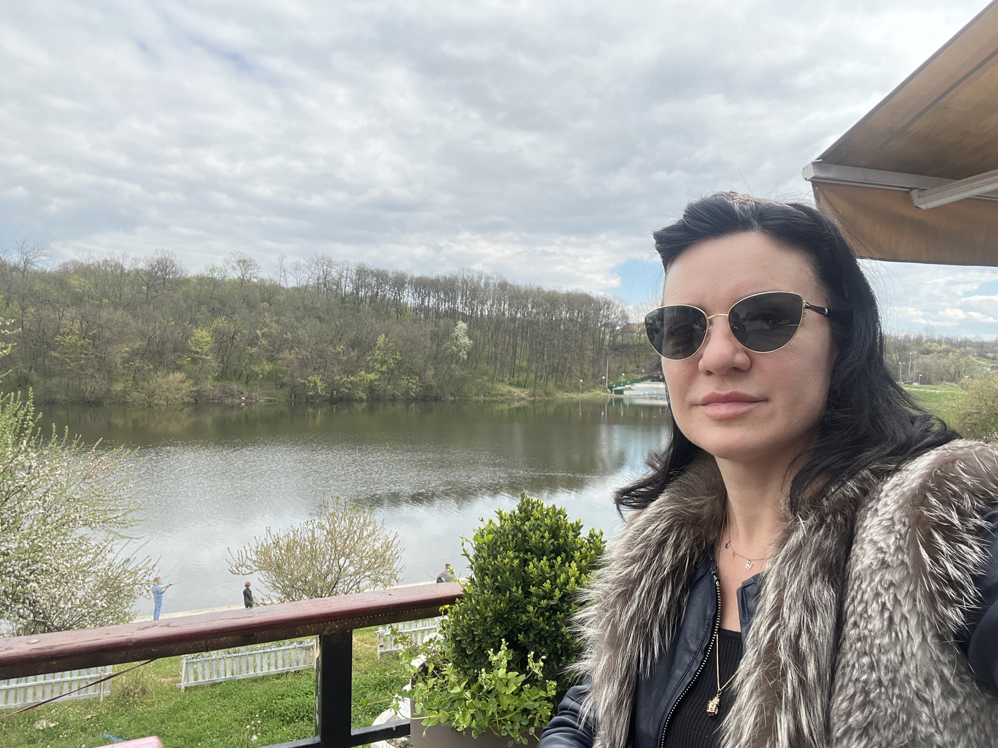 A woman wearing sunglasses and a fur-collared jacket is taking a selfie. She is on a balcony with a wooden railing. Behind her is a lake, with trees lining the opposite shore. Two figures are visible on the shoreline near the left edge of the frame, one is wearing a light blue outfit and the other is wearing black, and appear to be fishing. A white picket fence is visible below the railing. A potted plant is on the balcony. The sky is cloudy.
Without additional information, I cannot provide the exact location in Smederevska Palanka Municipality.