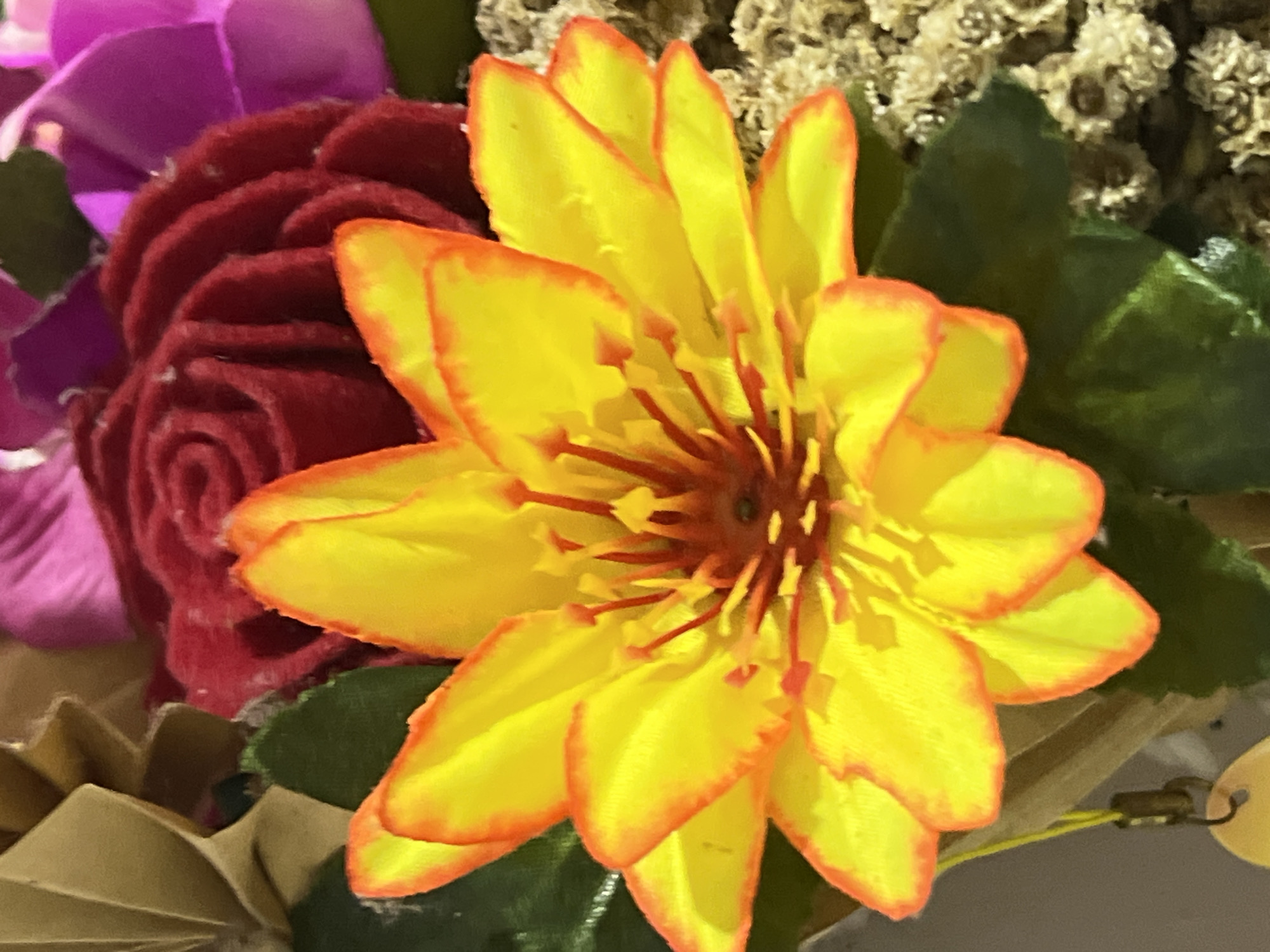 The image shows a close-up view of a bright yellow flower with orange-red edges, likely a lotus or dahlia, in the foreground. The flower has prominent stamens in the center. Behind it, there is a deep red, rose-like artificial flower and a hint of a bright pink artificial flower. The background is out of focus, with some greenery and what appears to be dried floral elements or a decorative basket. The lighting suggests an indoor setting, possibly a shop or home where artificial flowers are displayed or sold. There is no visible indication of the specific location within Denpasar, Indonesia, as the image lacks any recognizable landmarks or context specific to that city.