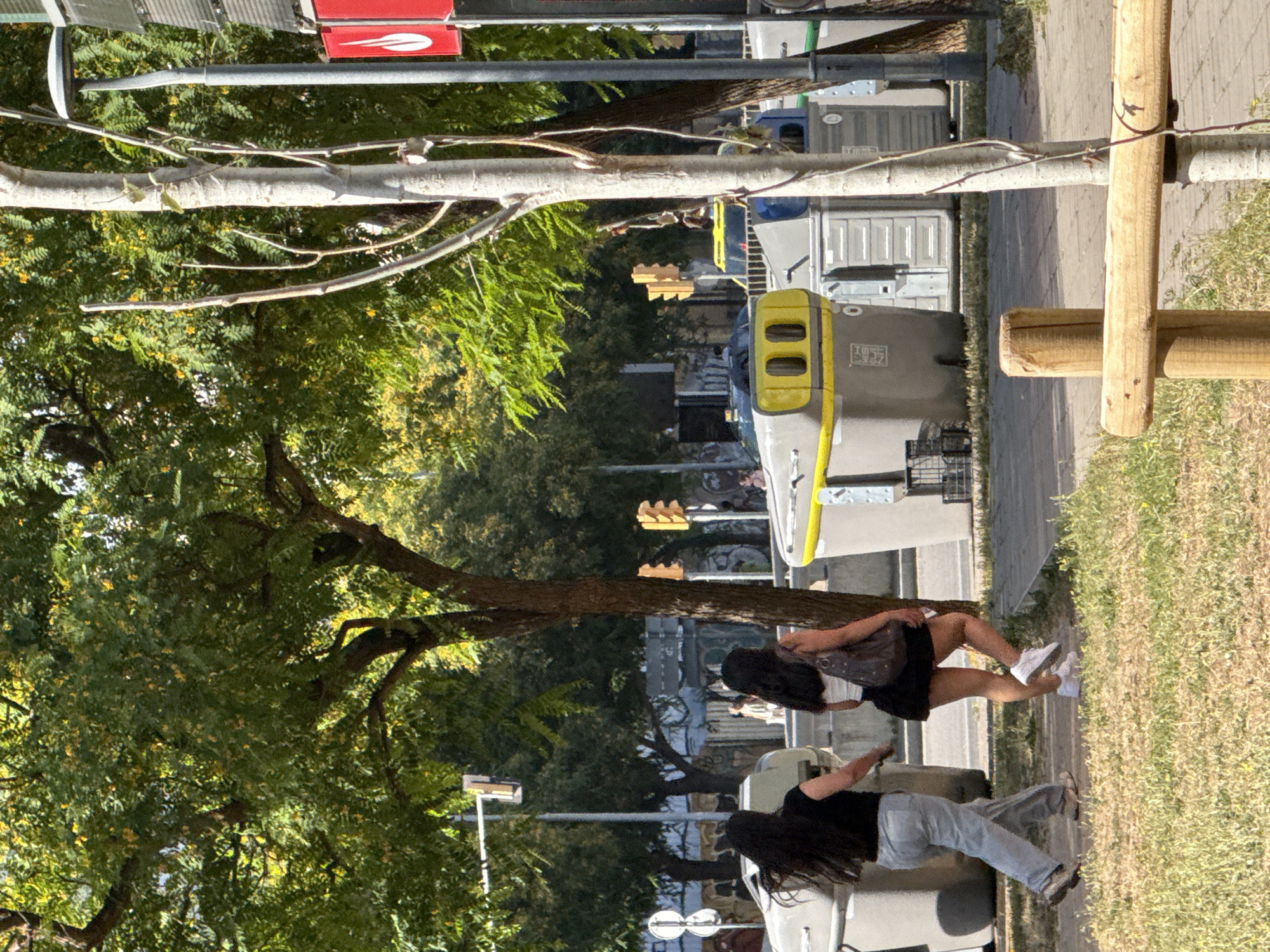 Two young women walk along a sidewalk in L'Hospitalet de Llobregat, Spain. The woman on the left has long dark hair and wears a black t-shirt and light grey sweatpants. She is walking away from the camera, and her left hand is visible. The woman on the right has long dark hair and wears a white top, a black skirt, and white sneakers. She carries a dark purse over her right shoulder and is walking away from the camera. They are walking past several large, gray and yellow public recycling bins. In the background, traffic lights are visible, along with trees and buildings. The scene is set on a sunny day, with dappled light filtering through the trees. The foreground shows a patch of dry grass and a wooden fence post.