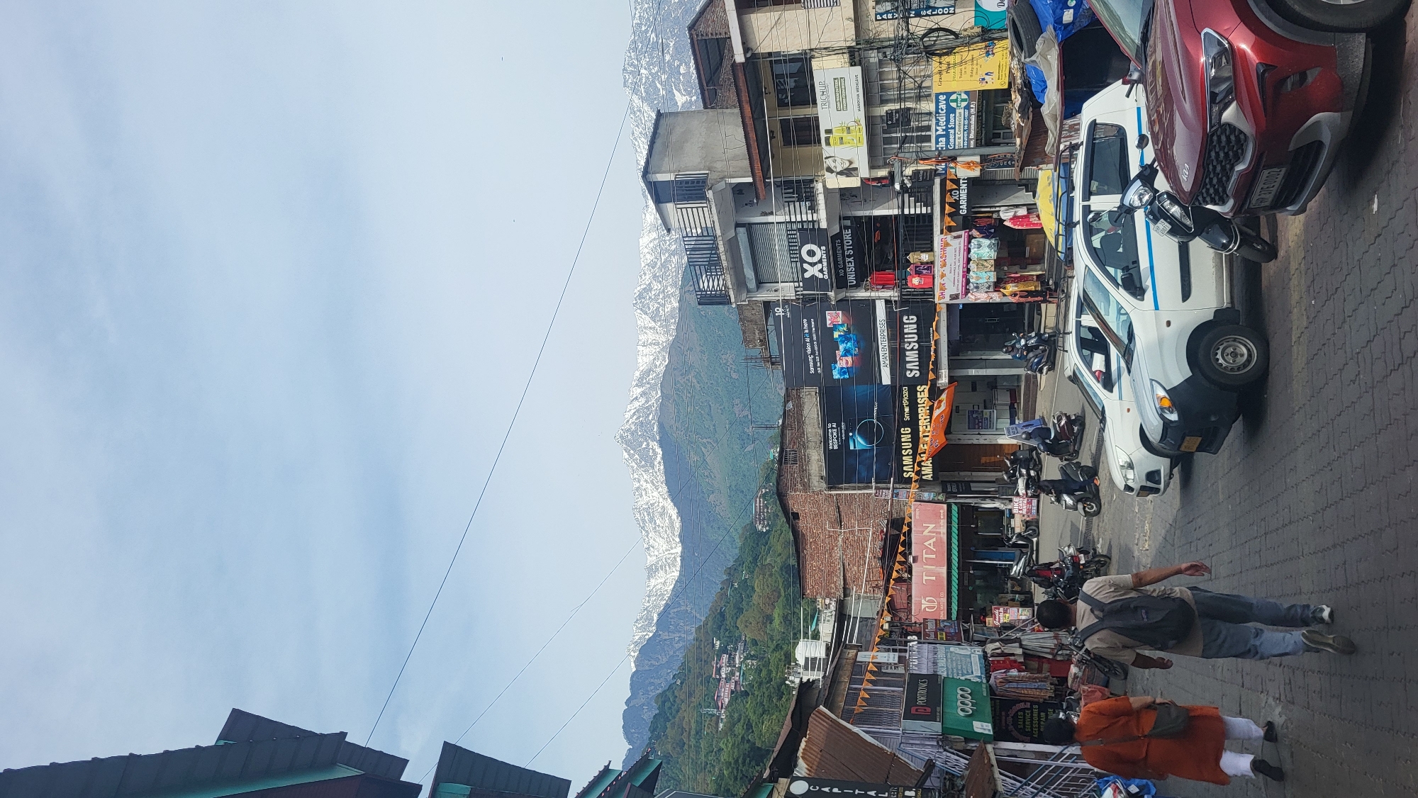 The image depicts a bustling street scene in Dharamshala, India, with a clear view of the snow-capped Dhauladhar mountain range in the background. Buildings line the right side of the frame, adorned with a variety of storefront signs for brands such as Samsung, Titan, Oppo, and XO Garments. Cars and motorbikes are parked haphazardly along the street. In the foreground, two pedestrians are visible – one in an orange garment, and another with a backpack. The sky is a pale blue, with the mountain range filling the space between the buildings and the sky.