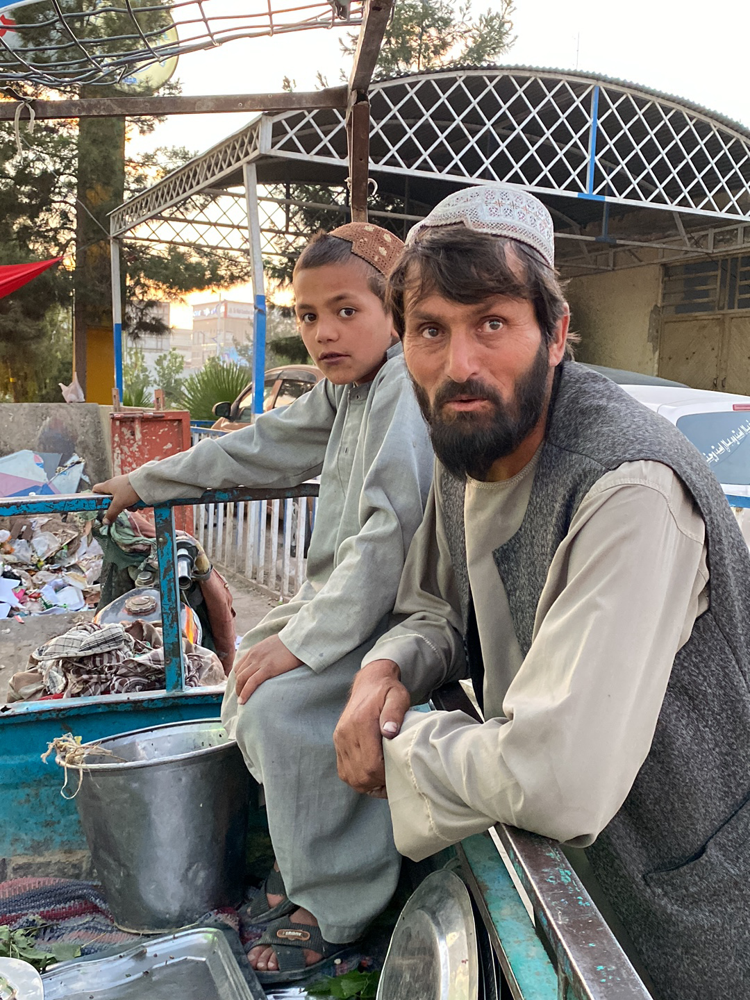 In a candid outdoor shot taken in the city of Kandahar, Afghanistan, a man and a boy sit close together. The man, likely in his late 30s or early 40s, sports a dark beard and a white skullcap, wearing a light-colored shirt with a dark vest. To his left, a young boy, wearing a brown skullcap and a matching light-colored outfit, gazes directly at the camera. Both are positioned within what seems to be the back of a small truck or cart, amidst various items including a large metal pot, a tray, and other miscellaneous objects. In the background, a building with a white, lattice-like awning can be seen, along with hints of trees and a partially visible vehicle. The lighting suggests that the photo was taken either in the early morning or late afternoon.